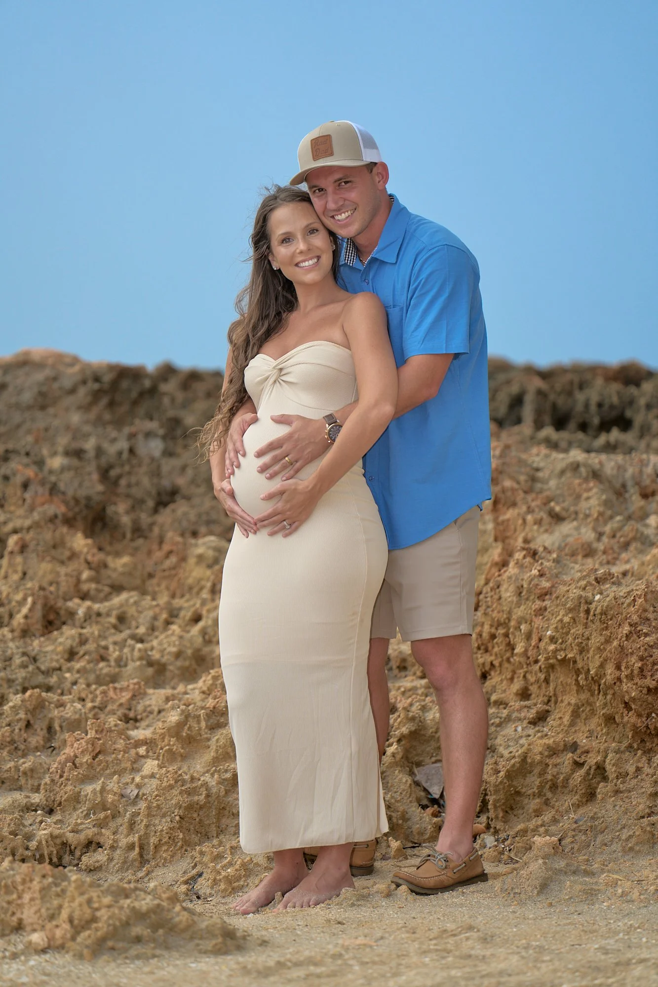 A happy pregnant woman and her partner standing on a rocky beach, with the woman wearing a beige strapless dress and the man in a blue shirt and shorts, both smiling.