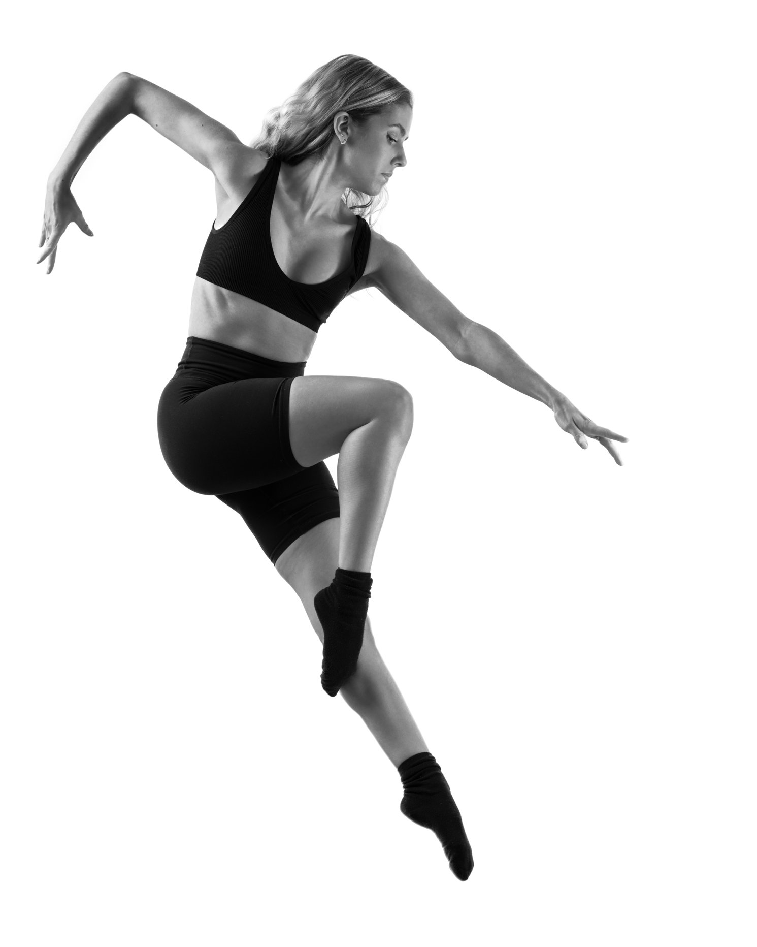Black and white photo of a female dancer mid-air, wearing athletic clothing, with one leg bent and the other extended, arms positioned gracefully.