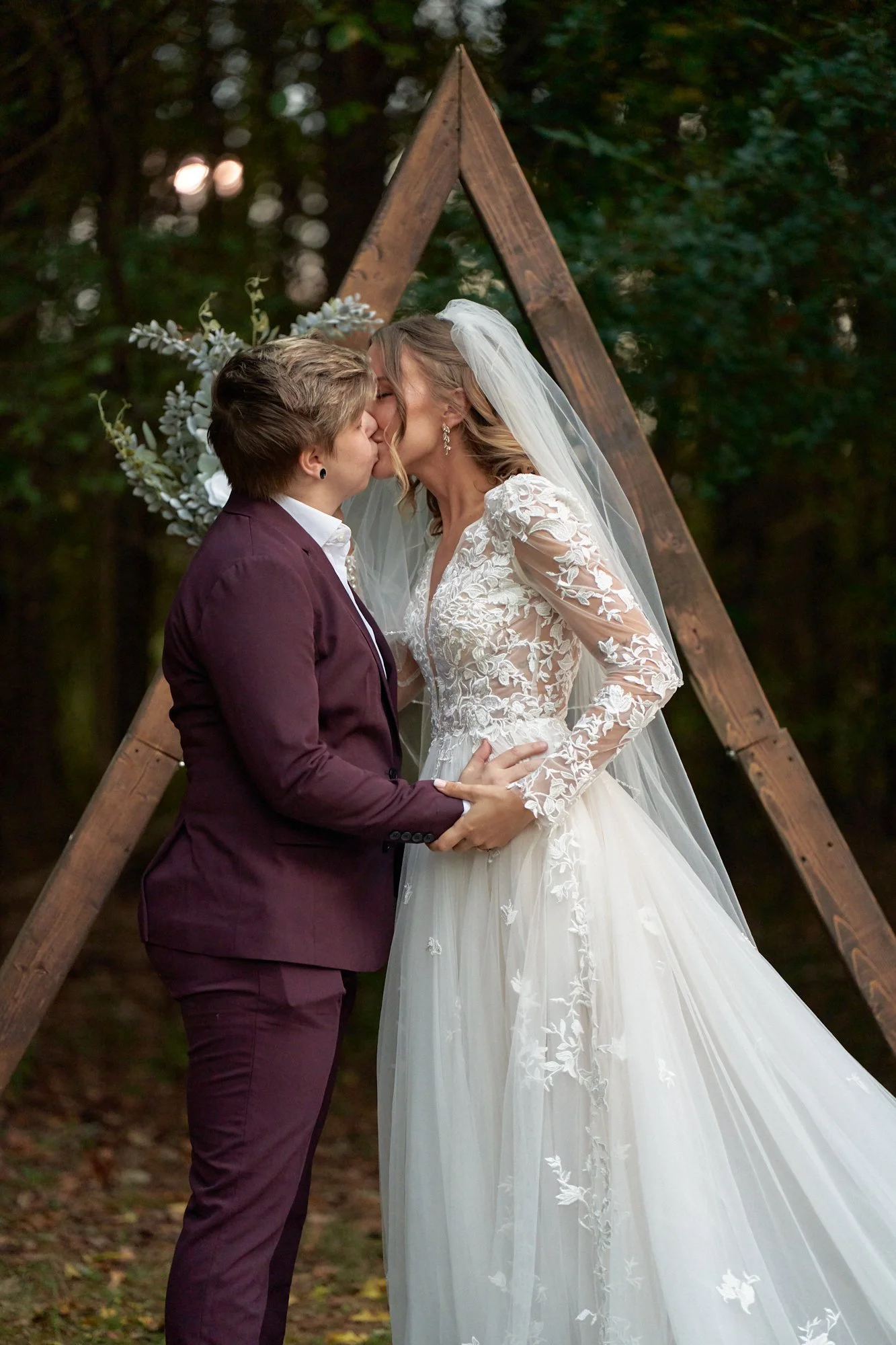 Two women kissing at a wedding ceremony outdoors, with a wooden triangular arch decorated with flowers and greenery behind them.