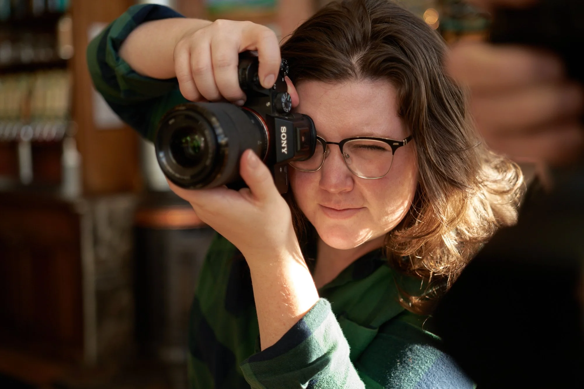 A woman with brown hair, glasses, and a green plaid shirt takes a photo with a black Sony camera, winking with her right eye, indoors with bookshelves in the background.