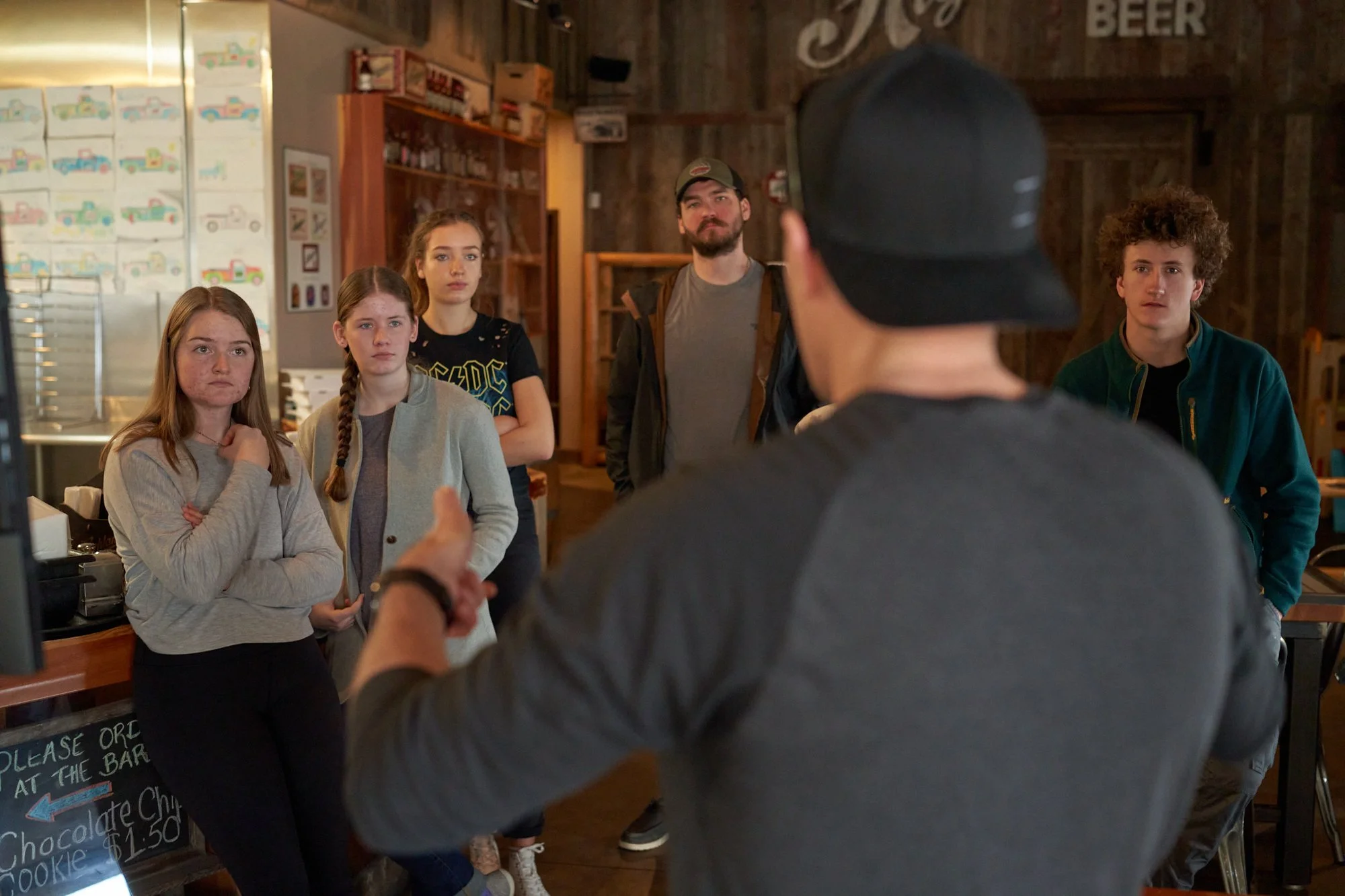 A man with a cap and gray shirt is speaking to a group of five teenagers in a rustic cafe or restaurant setting, with wooden walls and garage-style decor.