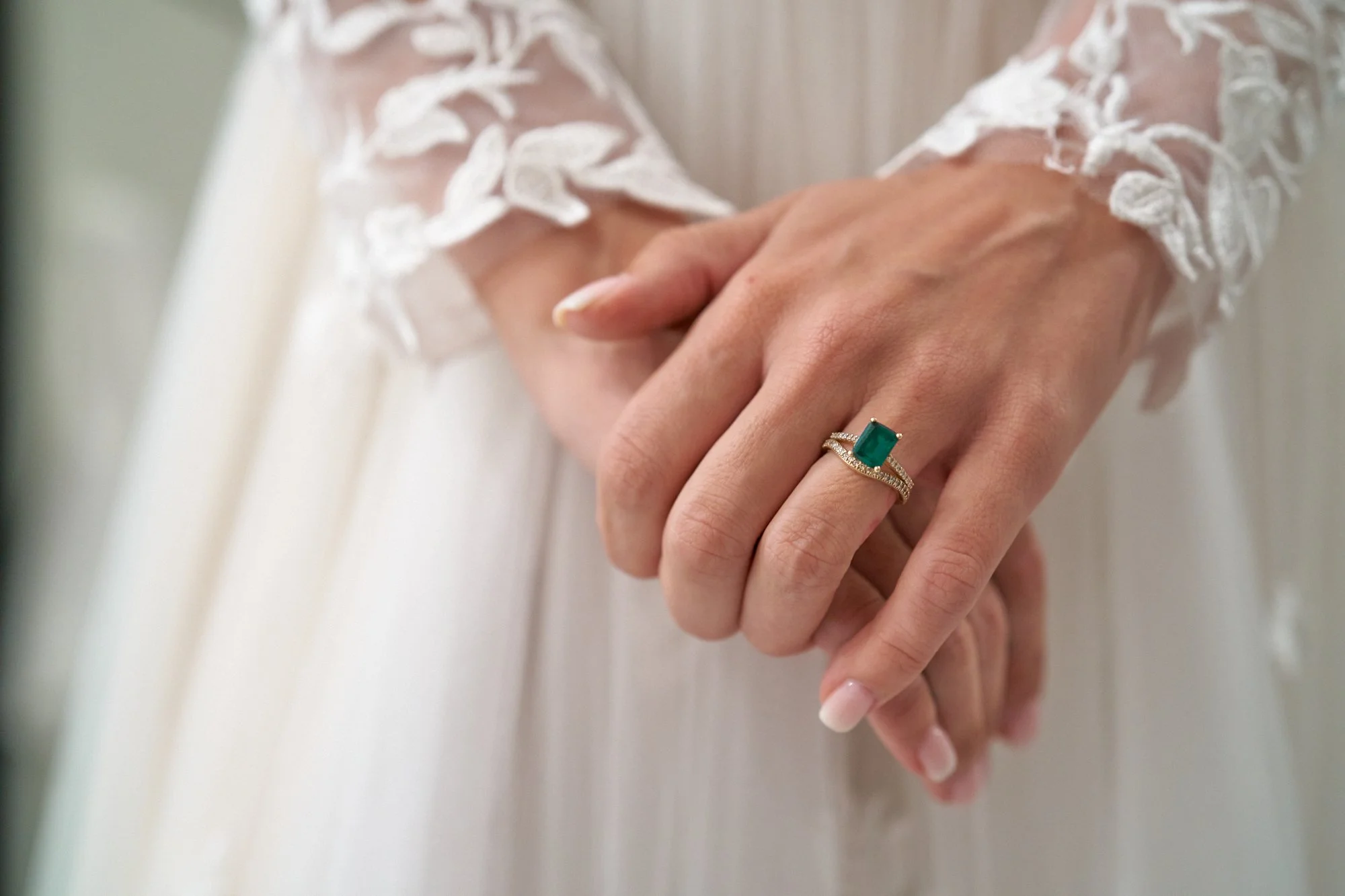 Close-up of a woman's hand wearing a green emerald ring with a diamond-encrusted band, resting on her other hand, with a lace sleeve visible.