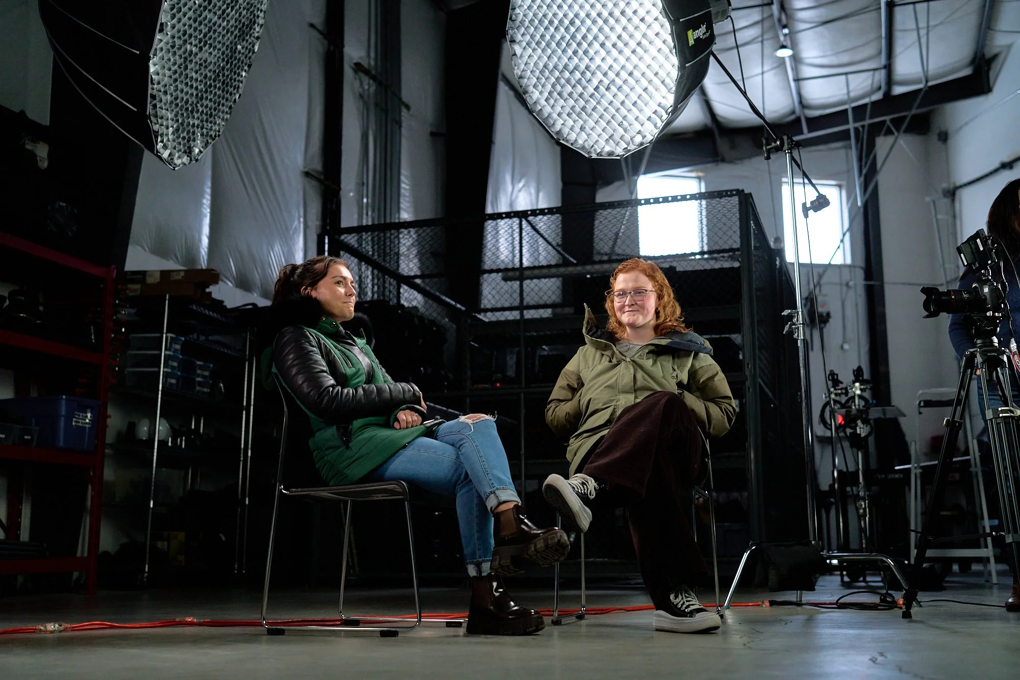 Two women sitting on chairs in a photography studio, surrounded by lighting equipment, with a camera on a tripod to the right.