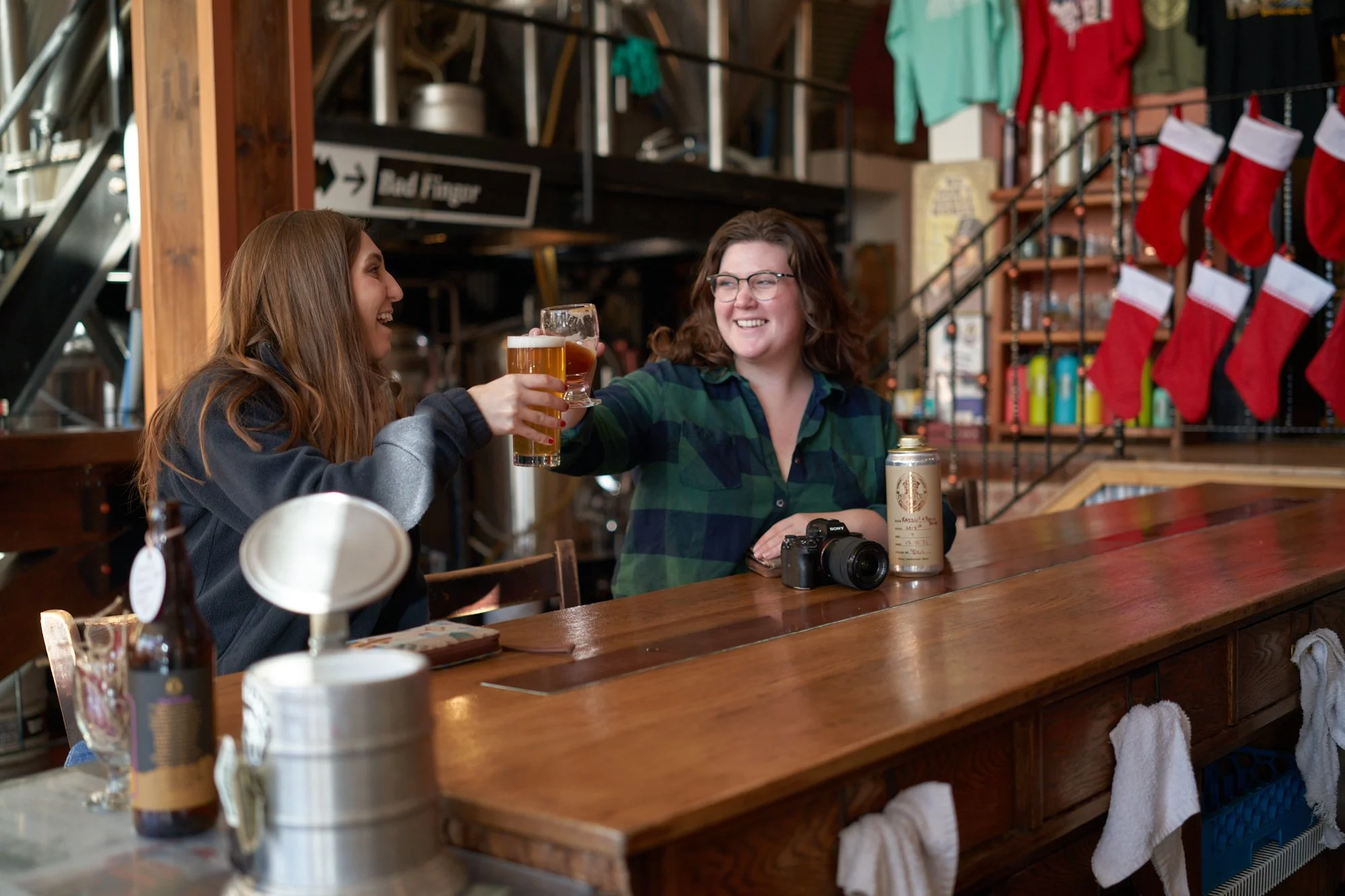 Two women sharing a toast with drinks at a bar, smiling and laughing, with Christmas stockings hanging on the wall behind them.