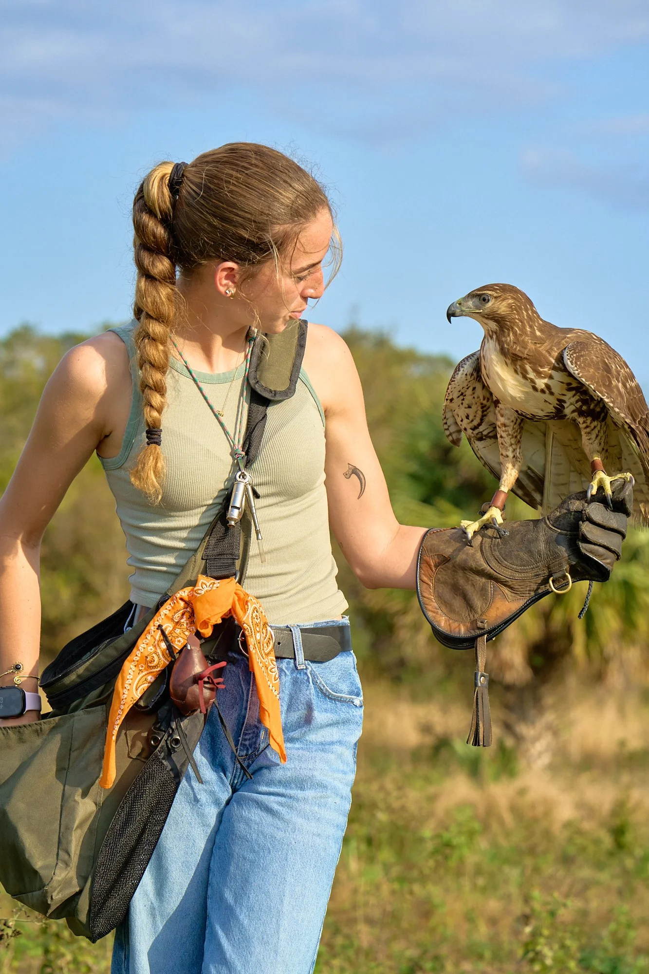 A young woman with a braid, wearing a sleeveless top and jeans, holding a hawk on her glove outdoors with a blue sky and greenery in the background.