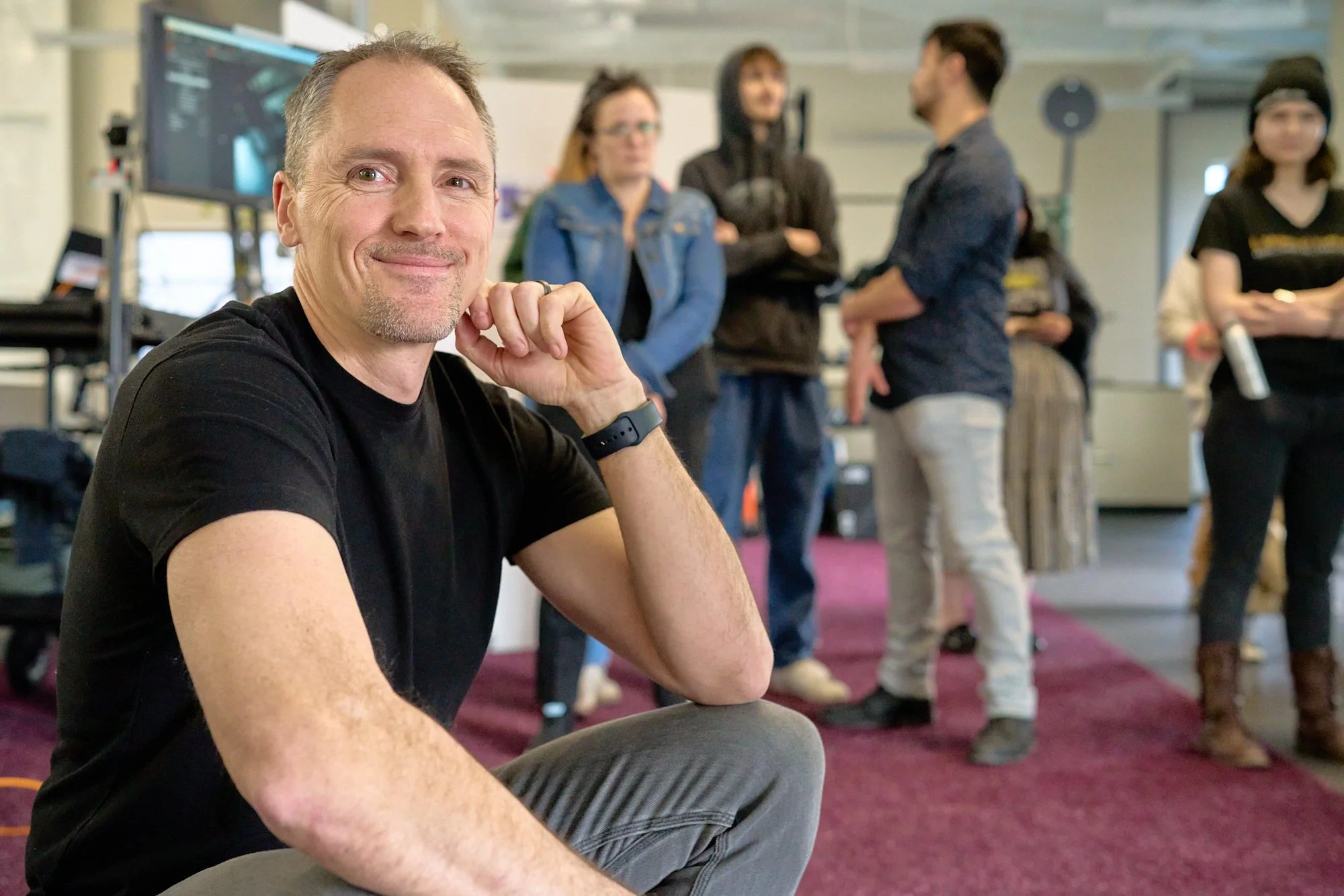 A smiling man in a black shirt sits on the floor in a room with a group of people in the background, some talking and others listening, with a large monitor on a stand and various equipment around.