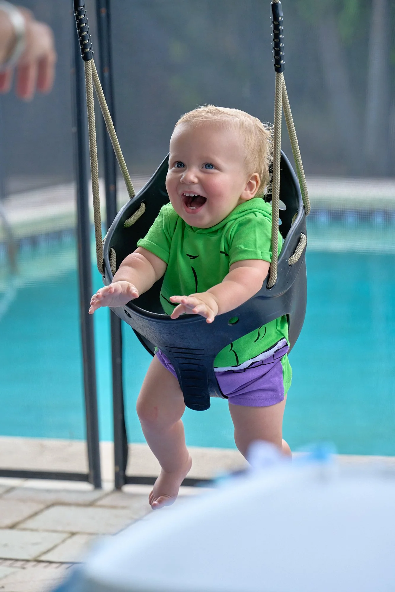 A young child with blond hair and a green shirt is happily swinging into a swimming pool in a baby swing seat, with a bright smile on their face.