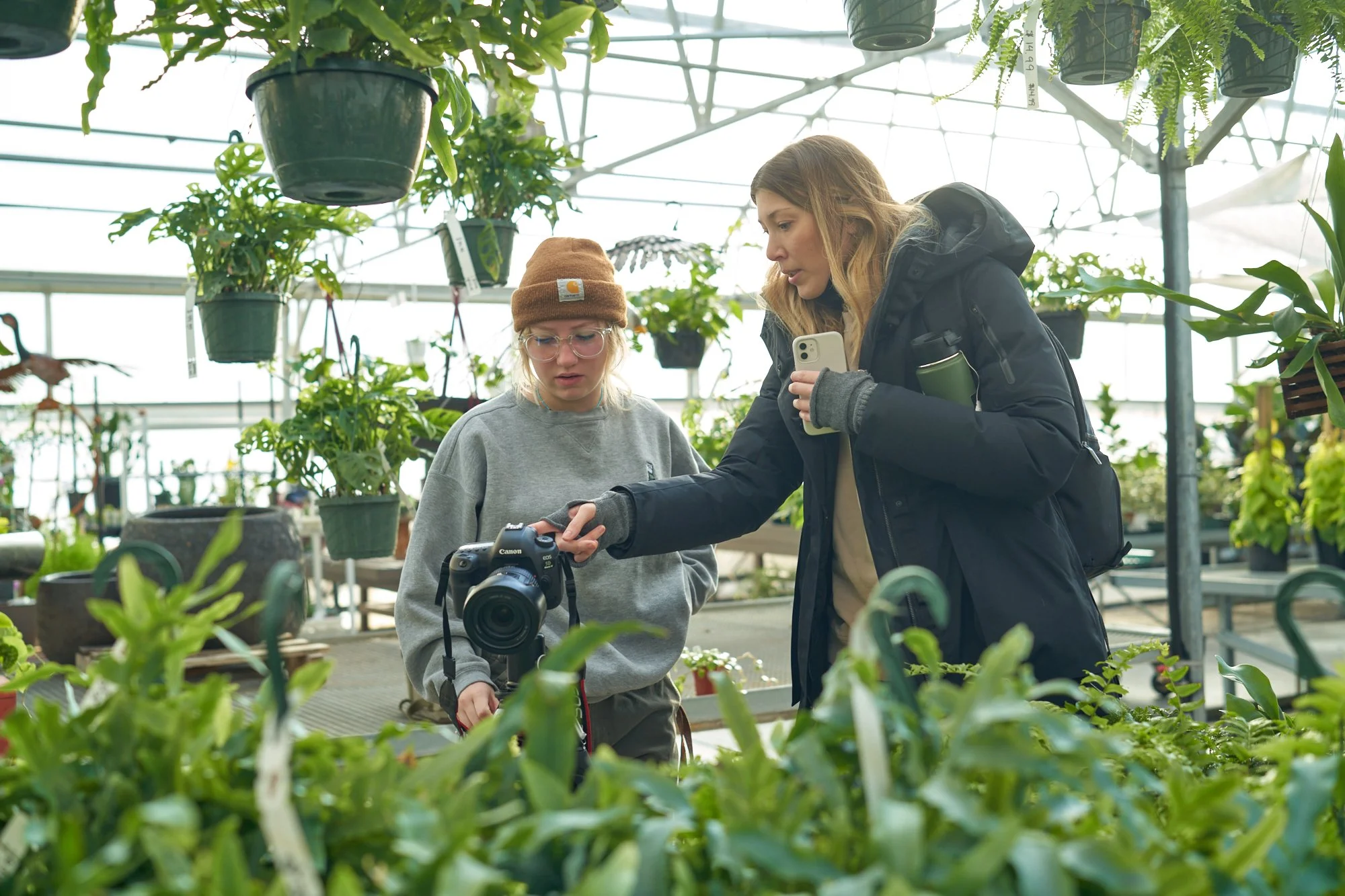 Two women are in a greenhouse surrounded by various green plants. One woman is holding a camera, while the other woman is pointing at something on the camera and holding a phone. They are engaged in photography or plant documentation.