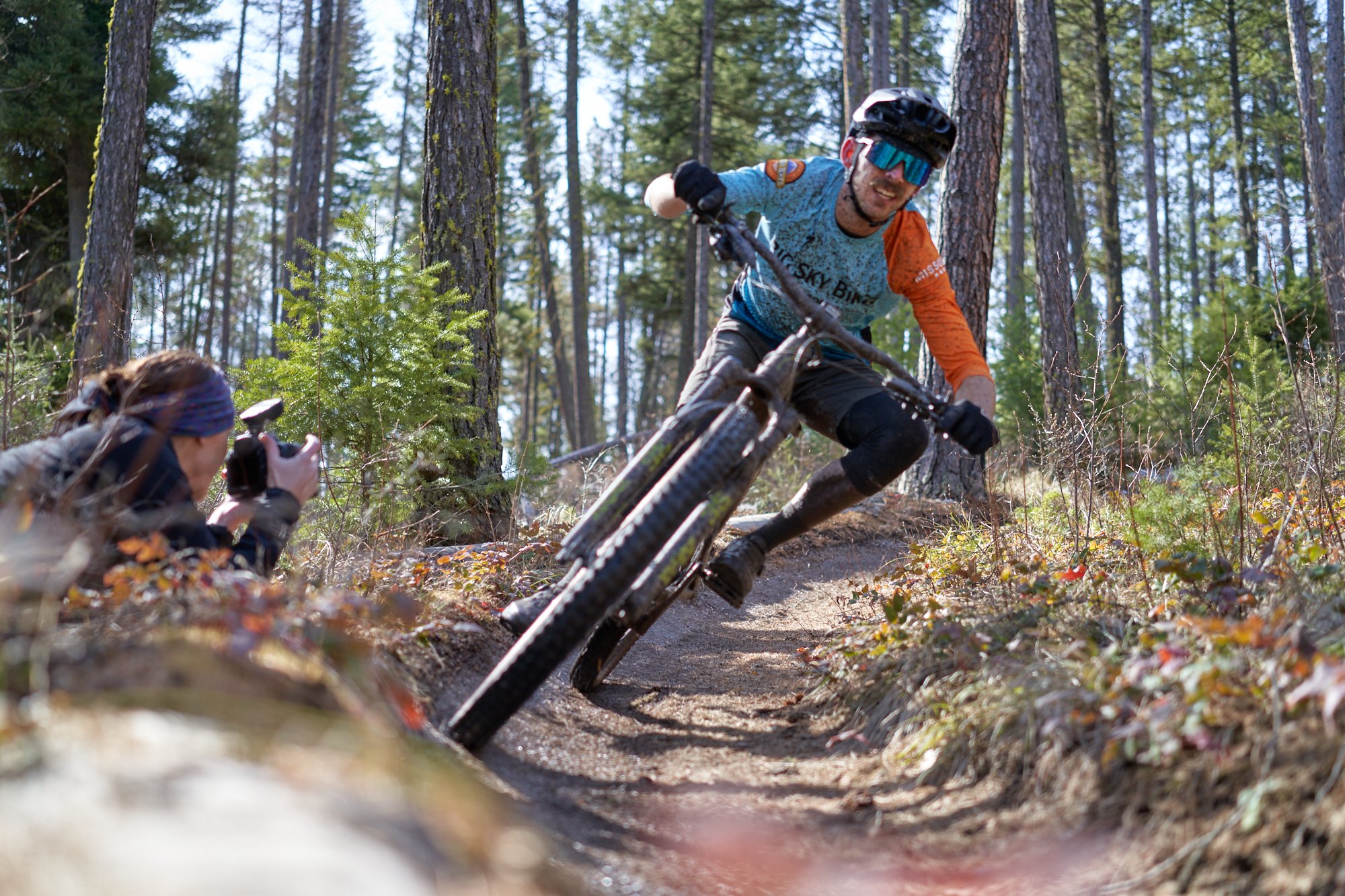 Mountain biker leaning into a turn on a forest trail, with a photographer crouched nearby taking pictures.