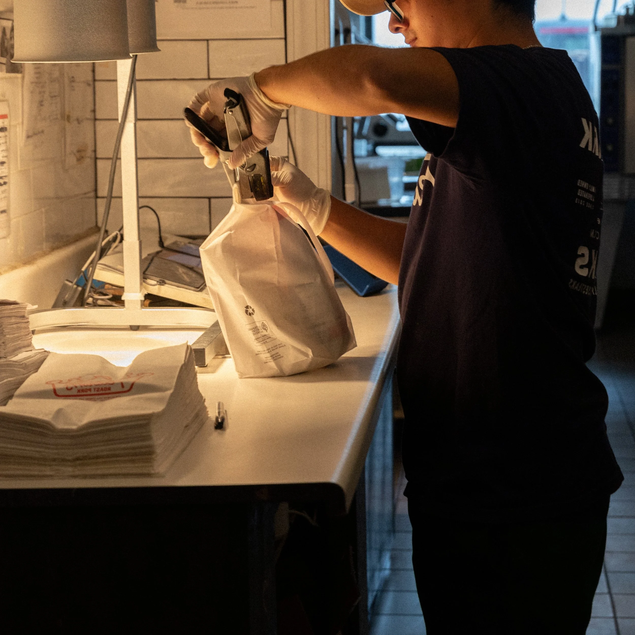 A person wearing glasses and gloves preparing food in a kitchen, filling a paper bag with an item.