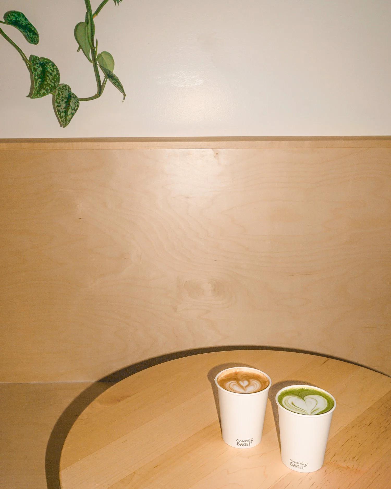 Two cups of latte art drinks on a round wooden table, one with a heart pattern in green and the other in brown, labeled 'Sincerely Bagel'.
