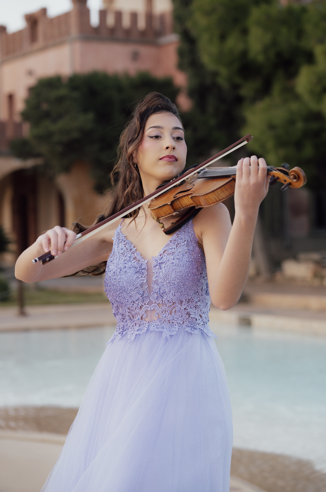 A young woman in a lavender lace dress playing the violin outdoors with a building and trees in the background.