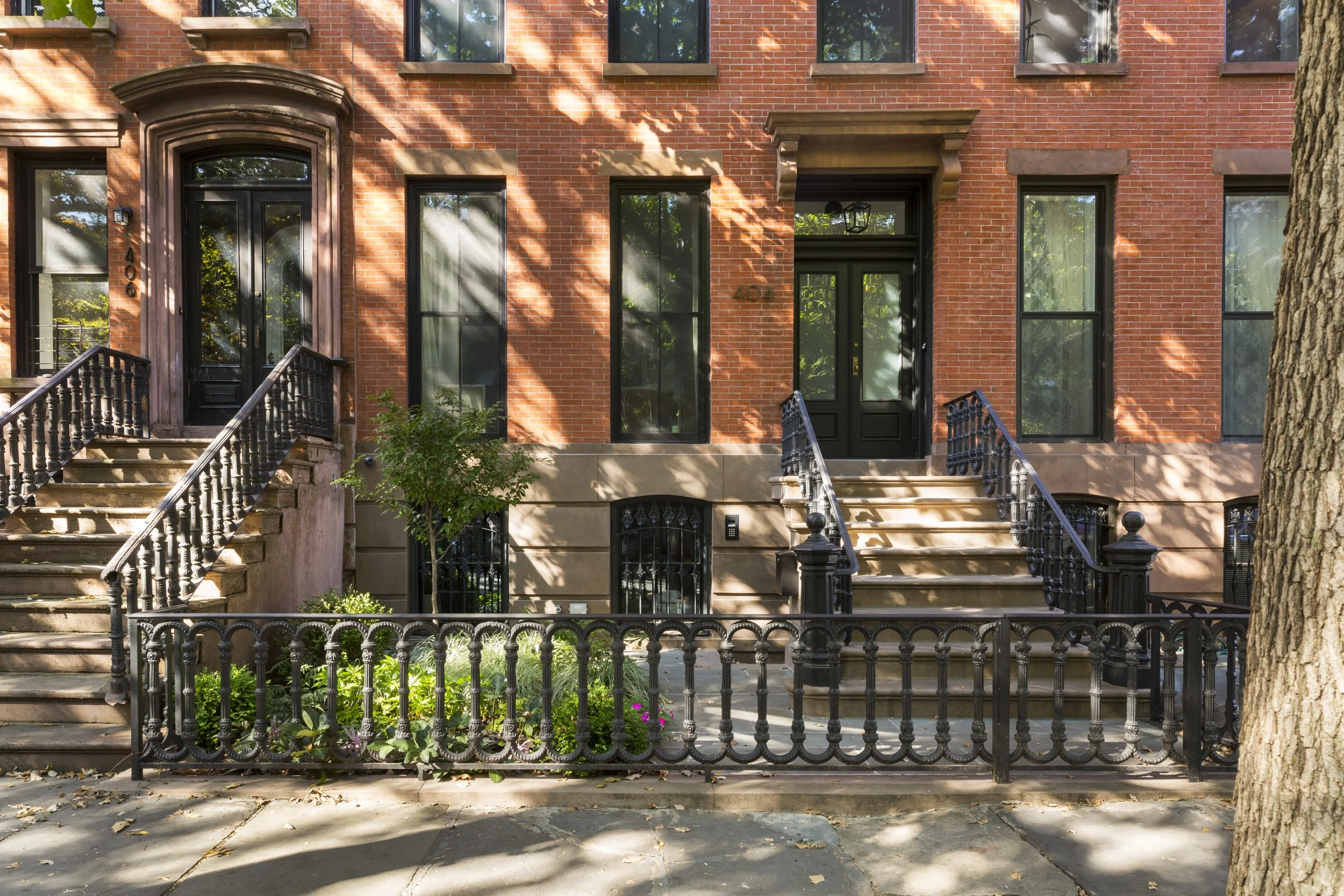 Brick townhouse with two black doors, staircase entrances, black iron railings, and a small front yard with plants, trees, and a sidewalk.
