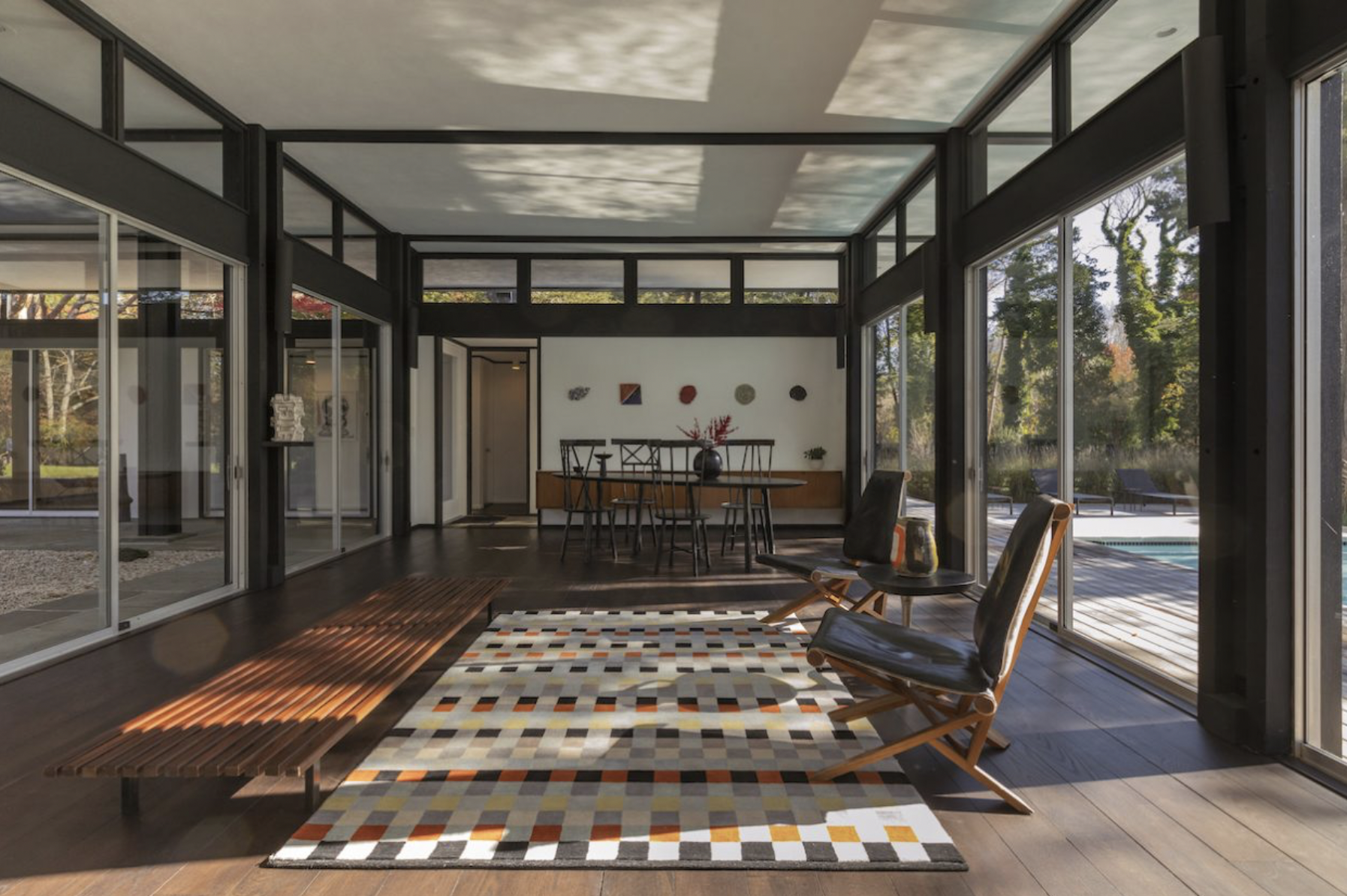 Sunlit modern living room with glass walls, black framing, wooden furniture, colorful geometric rug, black chairs, and artwork on the wall, with views of outdoor greenery and pool.