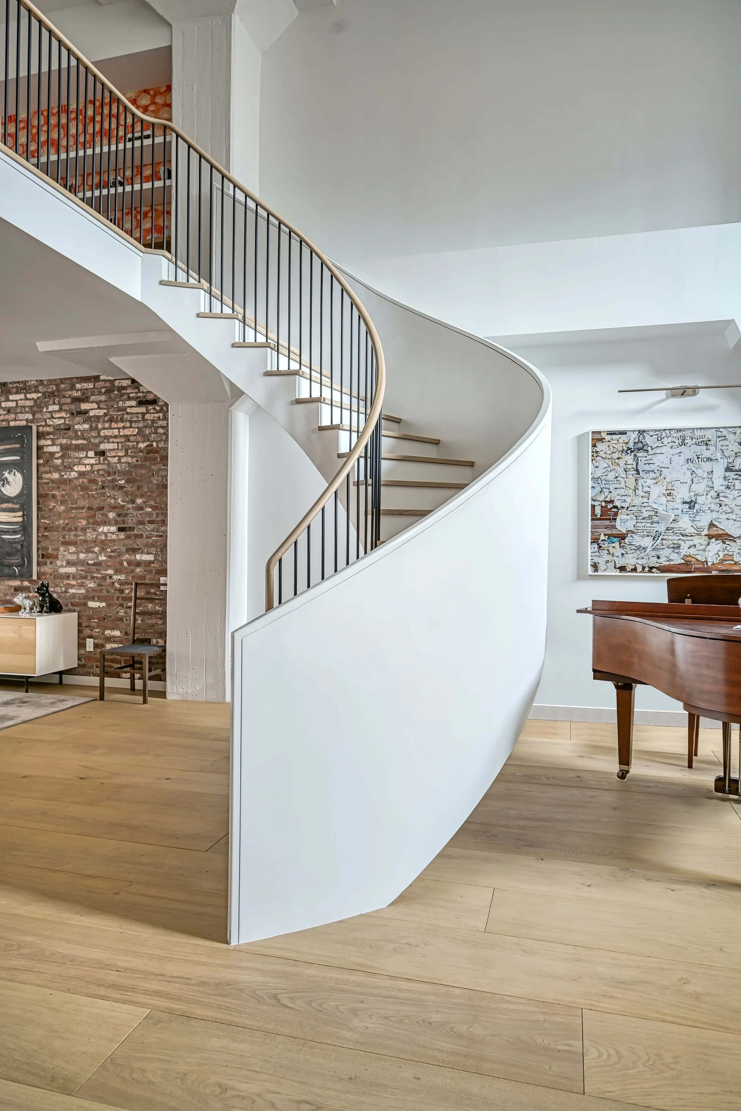 Interior of a modern house showcasing a curved staircase with black railings and wooden steps, a brick wall, a grand piano, and abstract wall art.