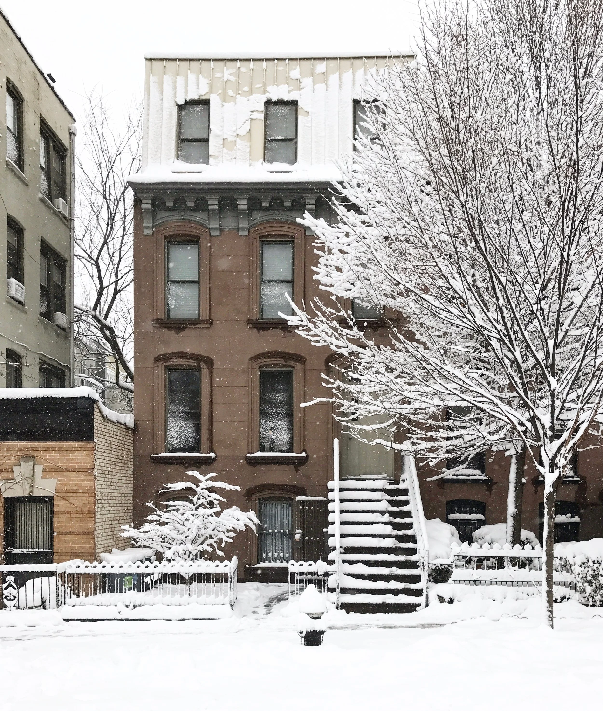 Brown brick townhouse with black stairs leading up to a second floor entrance, surrounded by snow-covered trees and ground in an urban neighborhood.