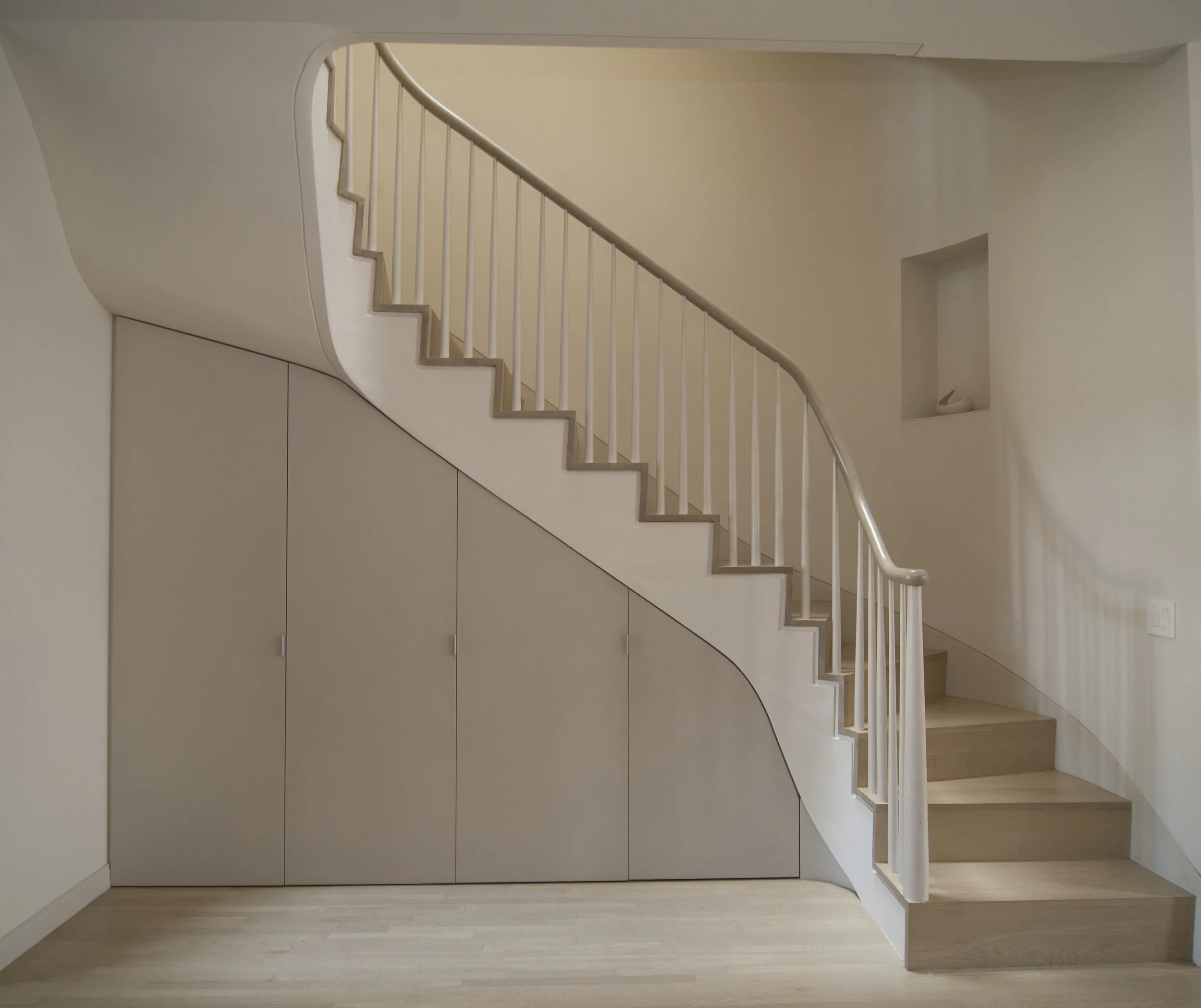 Interior view of a modern house showing a staircase with a curved balustrade and storage cabinets underneath, with a small niche on the wall.