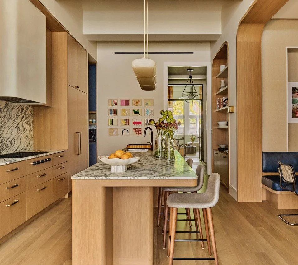 Modern kitchen with wooden cabinets, marble countertop, and apron sink. A rope light fixture hangs over the island, which has a bowl of oranges and a vase with flowers. There are three barstools with light-colored cushions at the island. A doorway in the background leads to a bright room with a window and a hanging geometric light fixture.