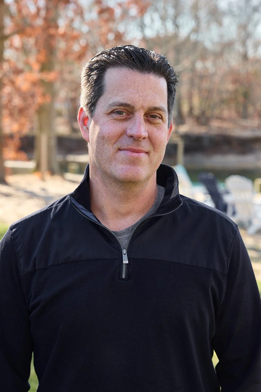 A man with dark hair slicked back, wearing a black zip-up jacket, standing outdoors with trees and lawn chairs in the background.
