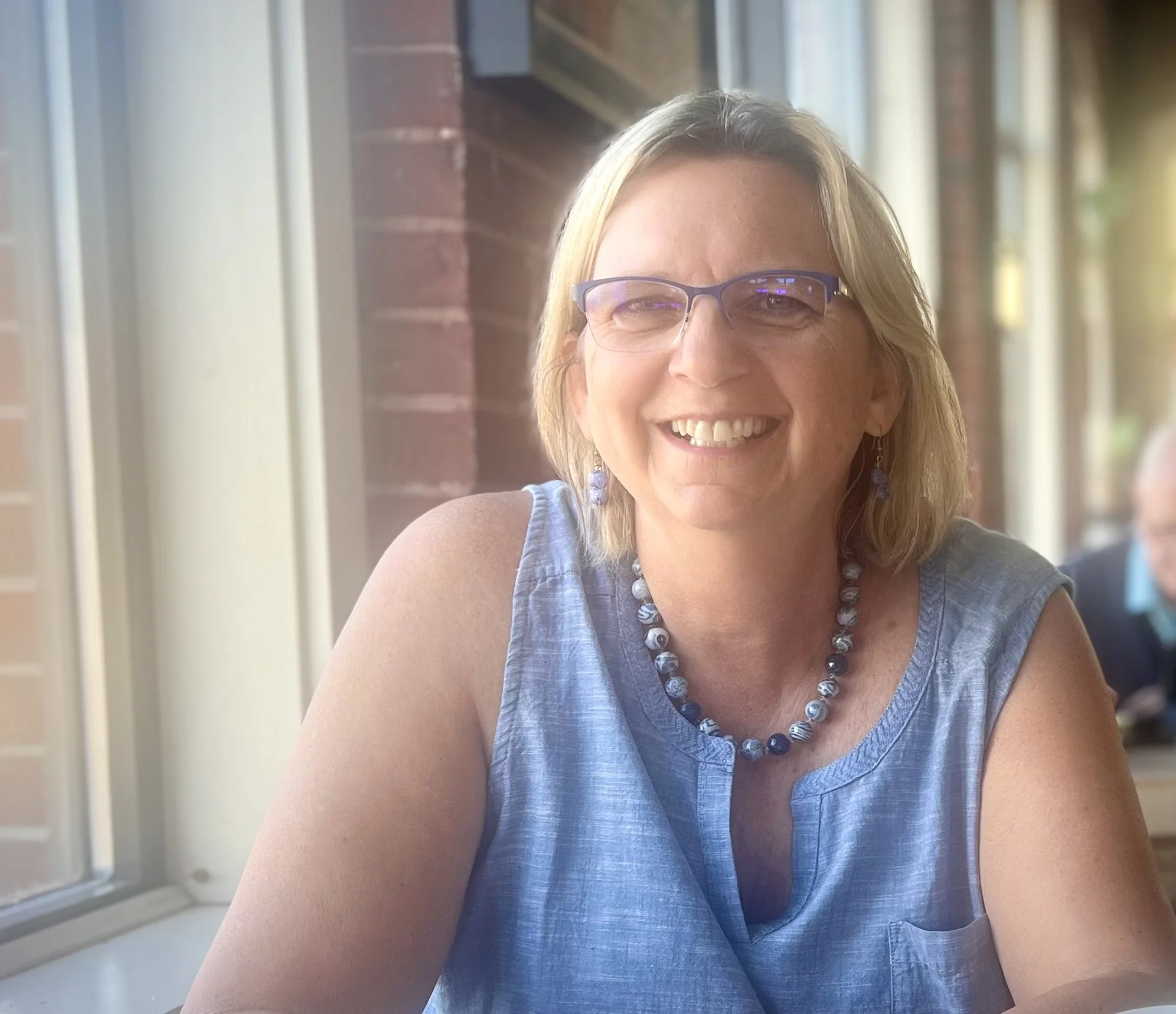 A smiling woman with blonde hair, wearing glasses, a blue sleeveless top, and a beaded necklace, sitting indoors near a window with natural light