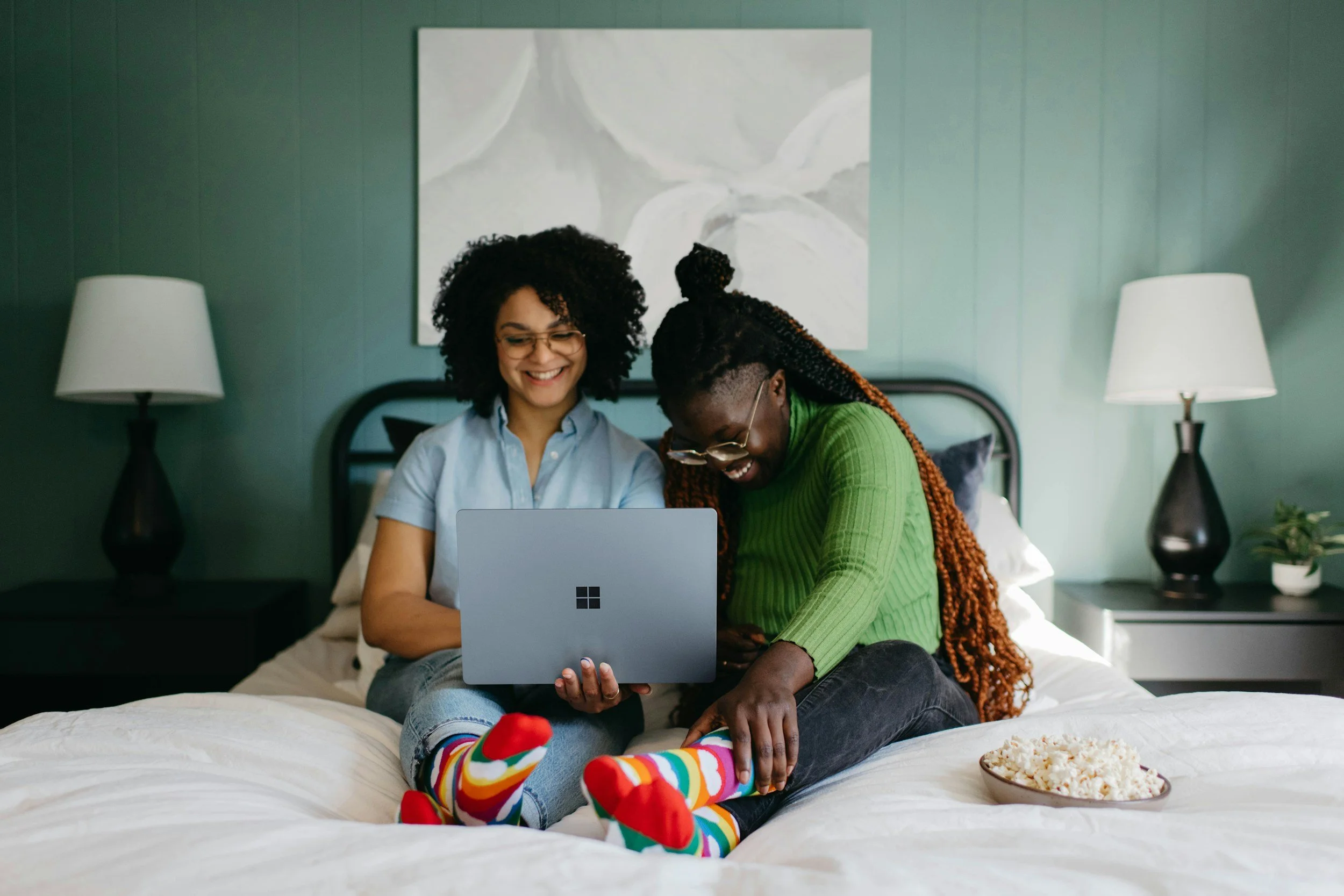 Two women sitting on a bed, looking at a laptop screen, smiling and laughing, with colorful socks and a bowl of popcorn nearby.
