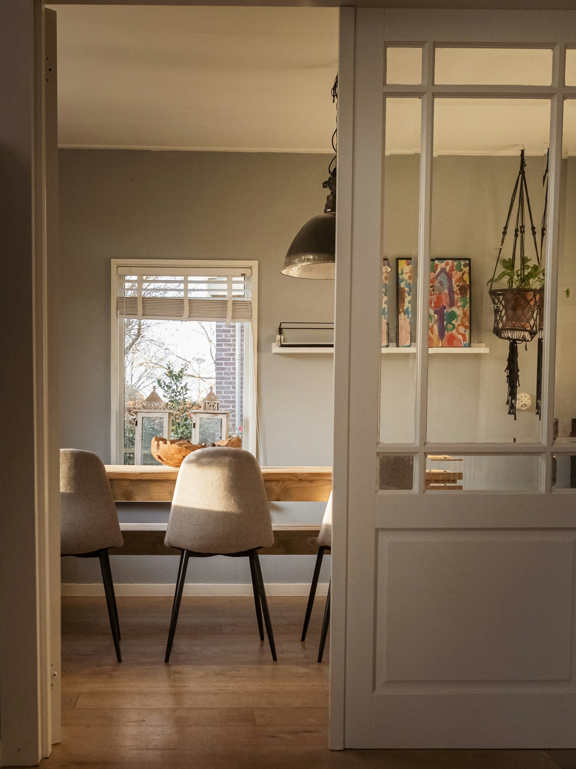 View of a cozy dining room seen through a door with glass panes, including a wooden table, beige chairs, a window with blinds, artwork on the wall, hanging plants, and warm lighting.