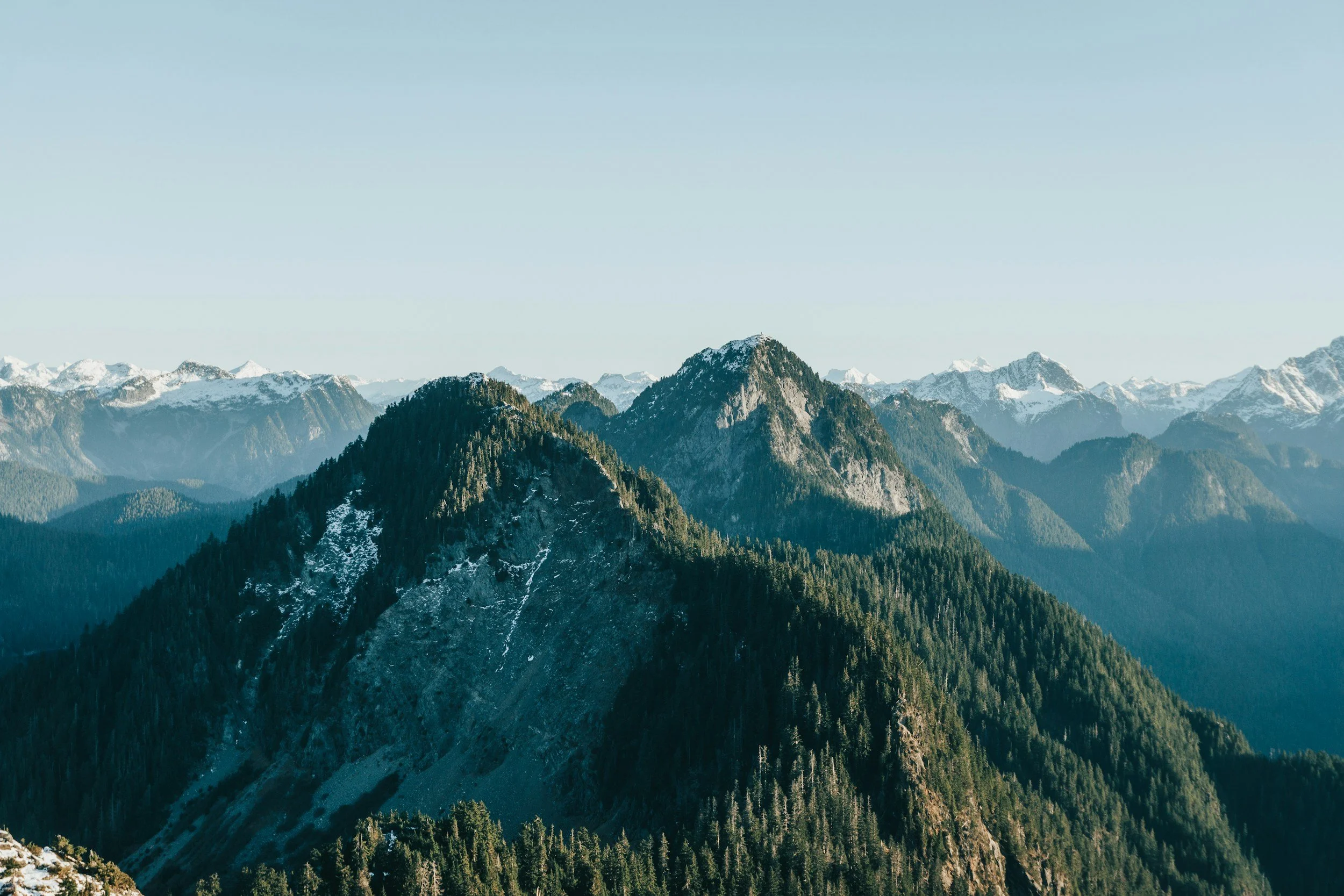 Scenic view of rugged mountain peaks covered with dense forests, with some snow patches, under a clear blue sky.