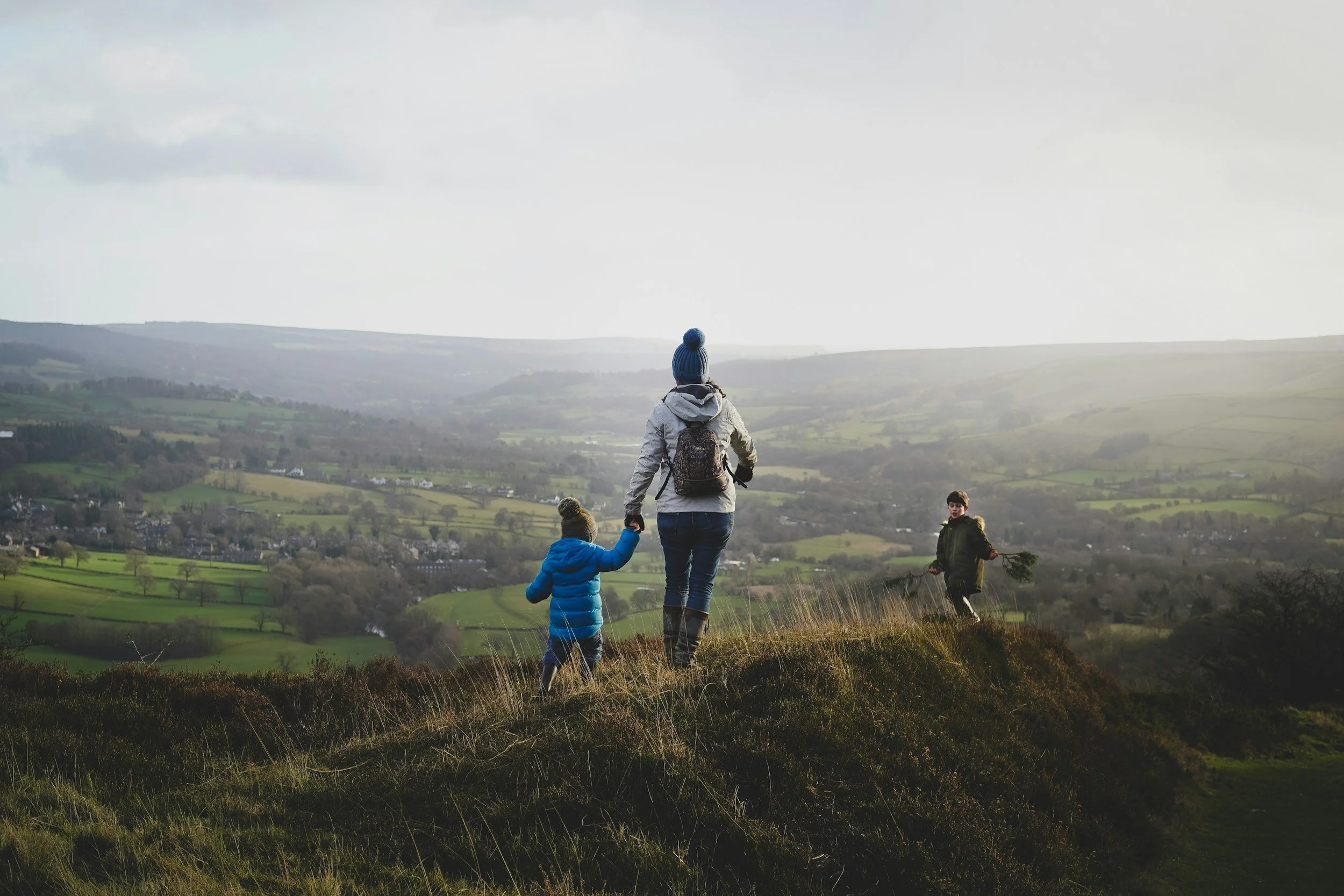 A woman walking with two children on a hillside overlooking a valley with green fields and scattered houses.