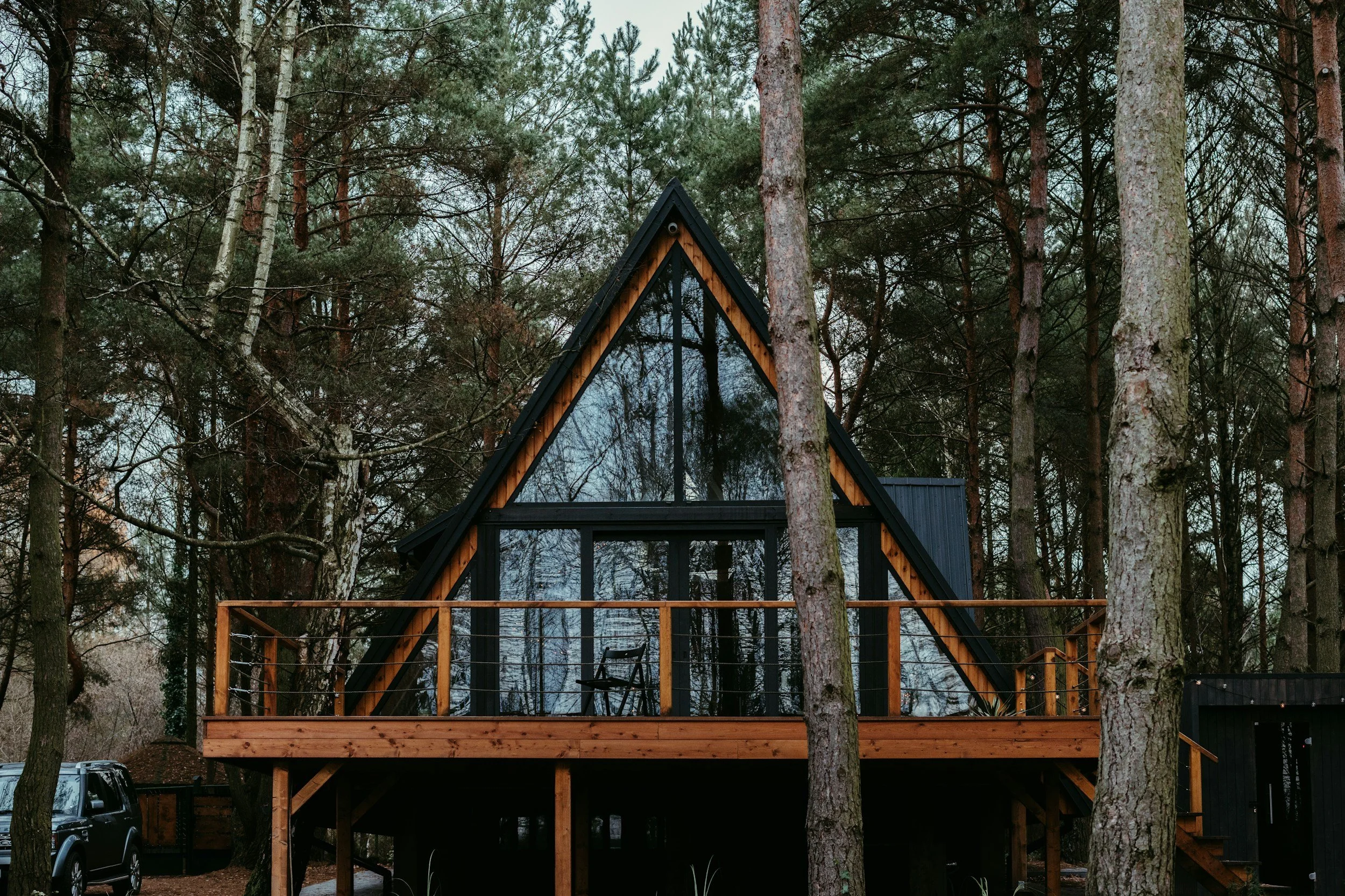 A modern A-frame house with large glass windows, on a wooden deck, surrounded by tall trees in a Canadian forest.