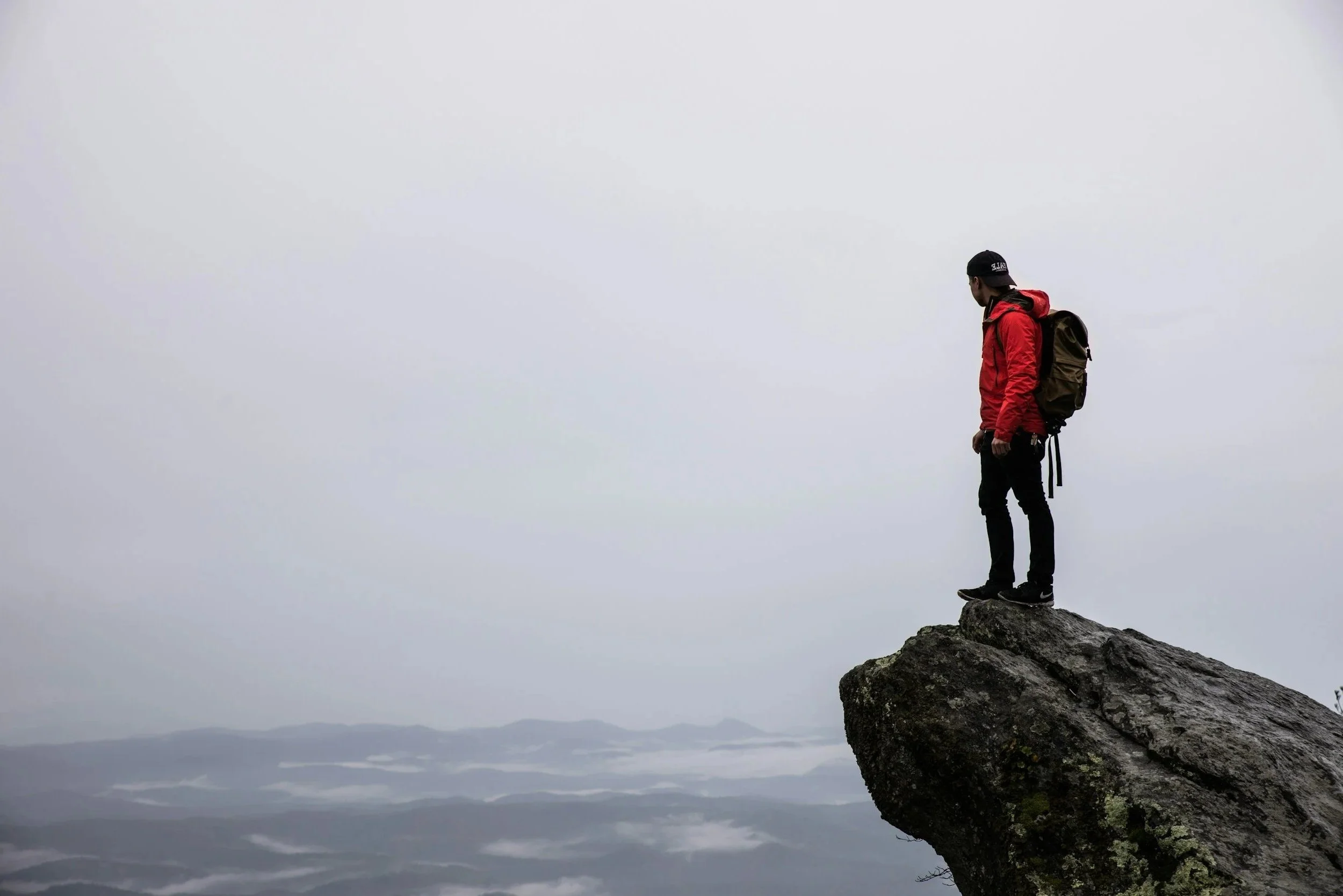 A person standing on the edge of a large rock overlooking a foggy mountain landscape.