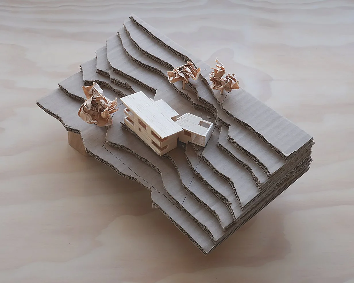 A miniature architectural model with layered cardboard representing a landscape, a small wooden building, and paper trees on a light wooden surface.