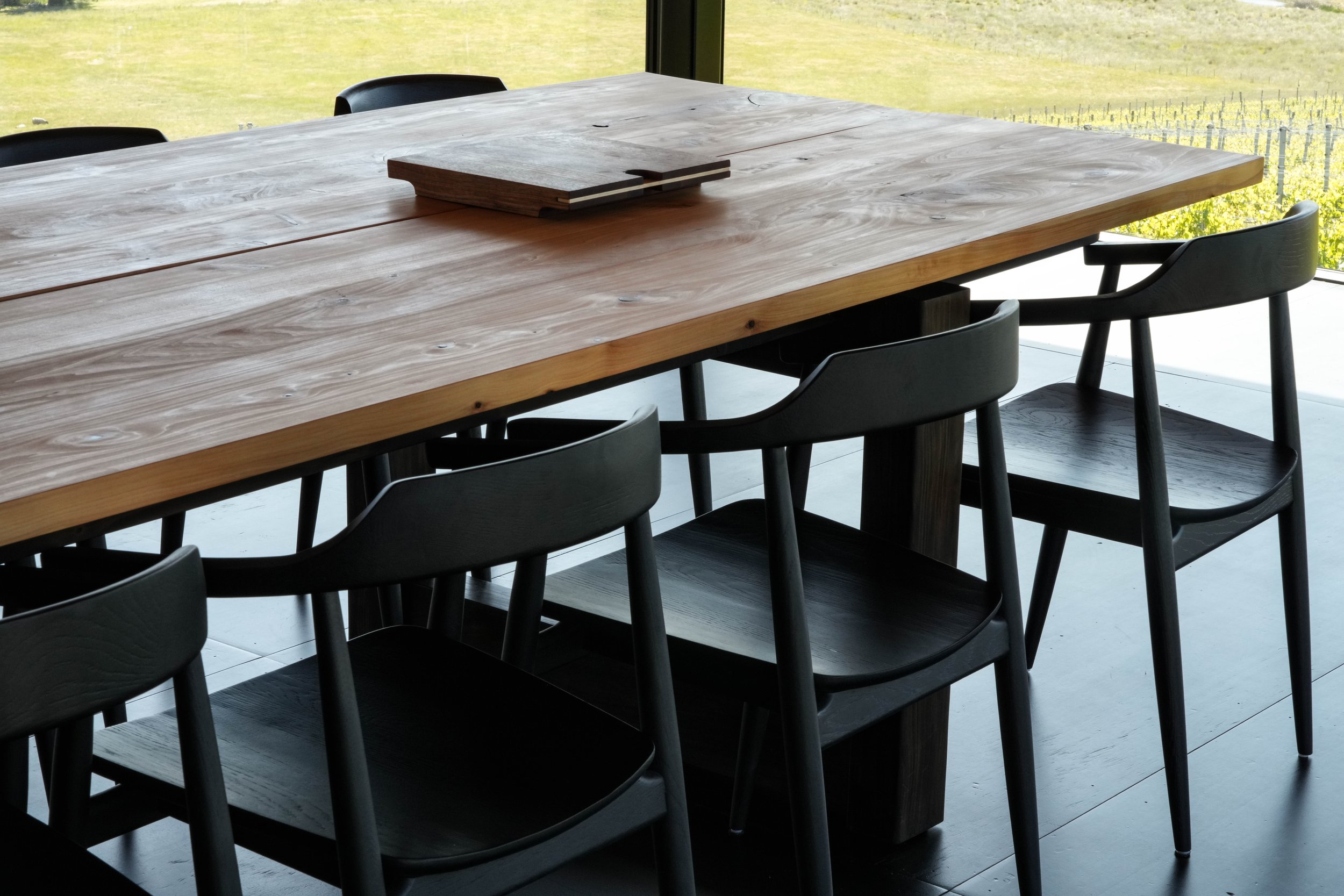 A large wooden designer dining table by Summa Studio with several black chairs around it, set in a room with large windows overlooking a beautiful landscape.