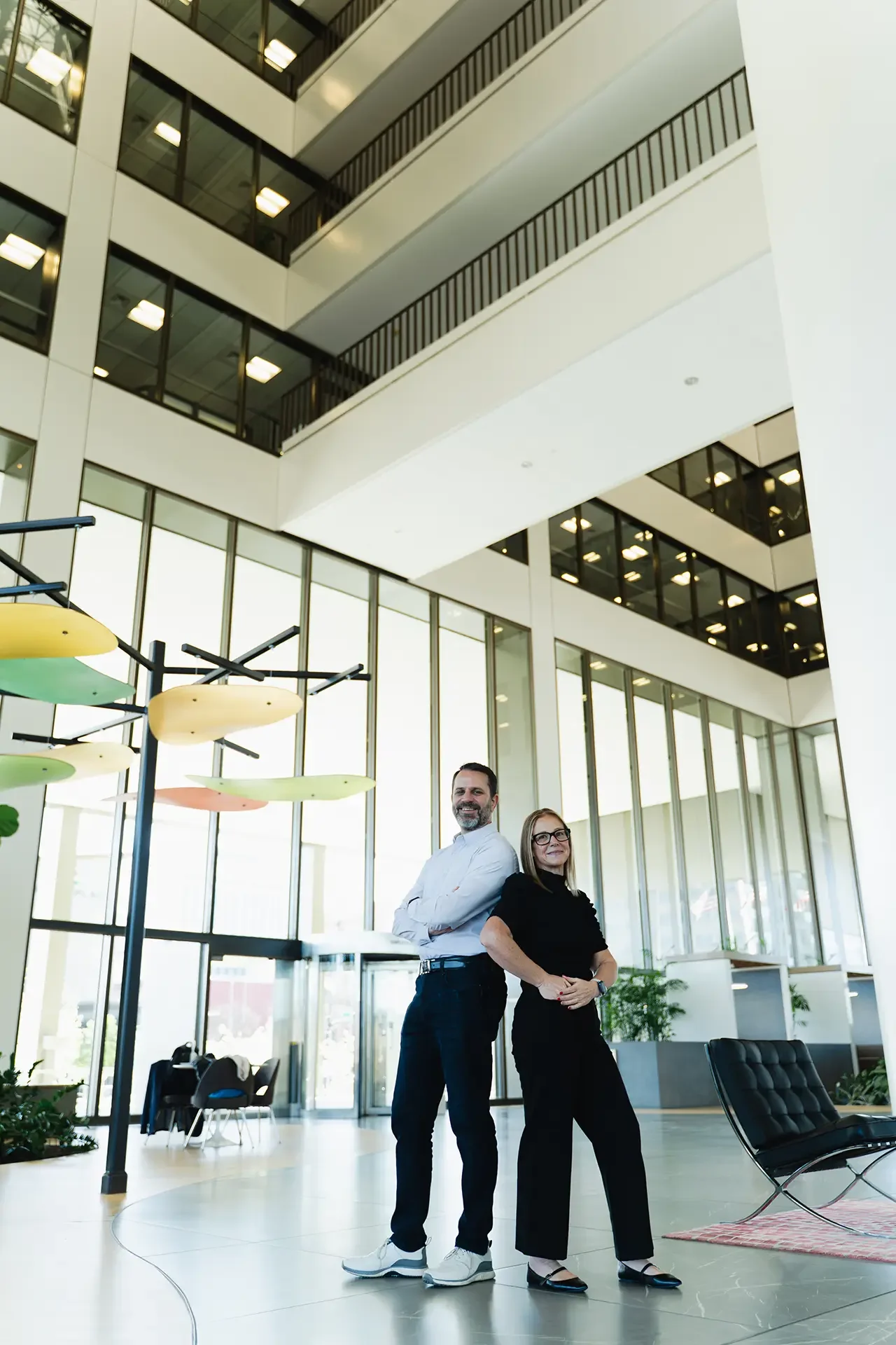 Two professionals, a man and a woman, stand confidently back-to-back inside a modern office building with large windows, multiple floors, and natural light.