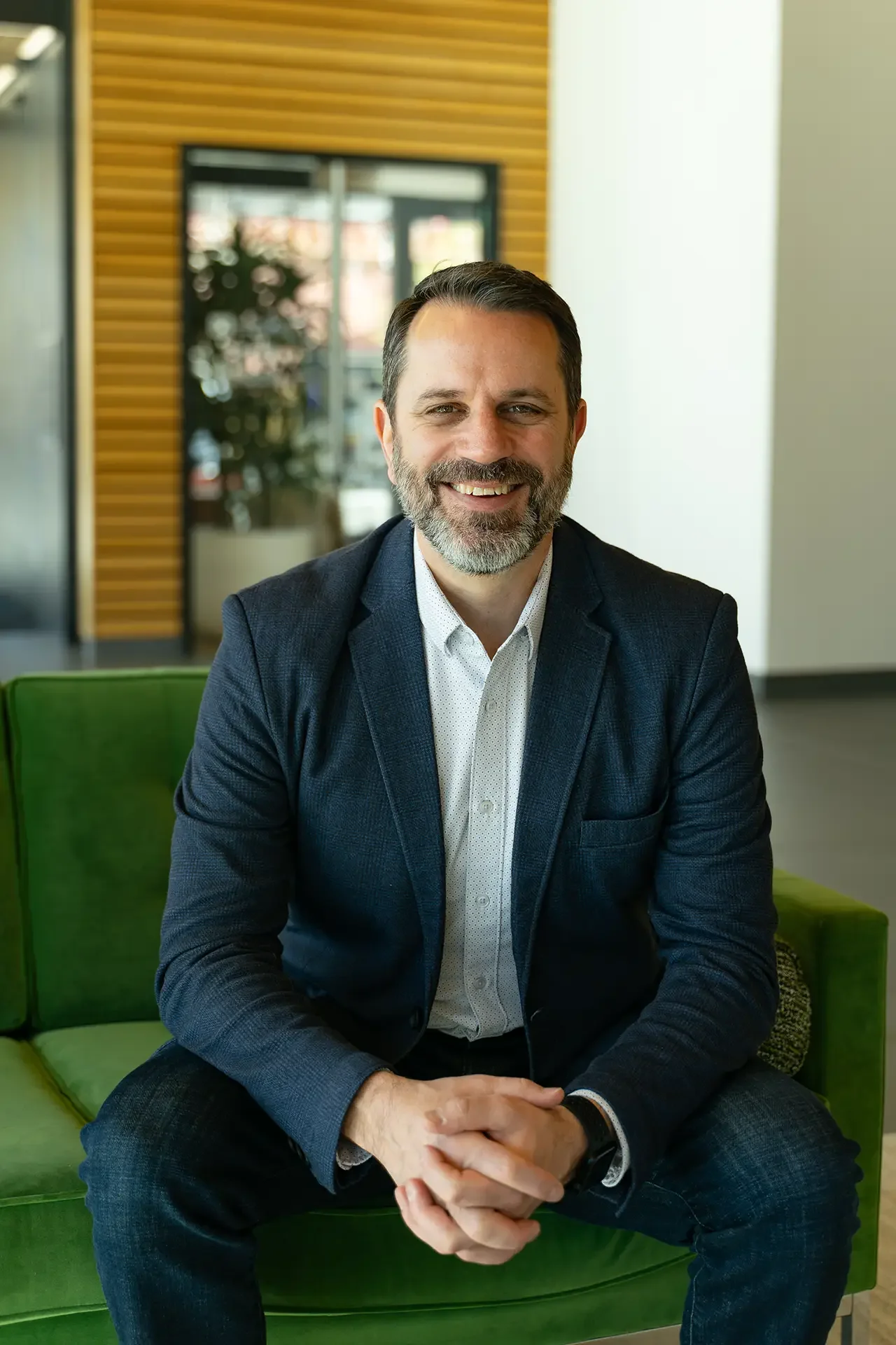 A smiling man with dark hair, a beard, and mustache, wearing a checked blue blazer over a white shirt, sitting on a green chair in a modern indoor setting with wood paneling and glass windows in the background.