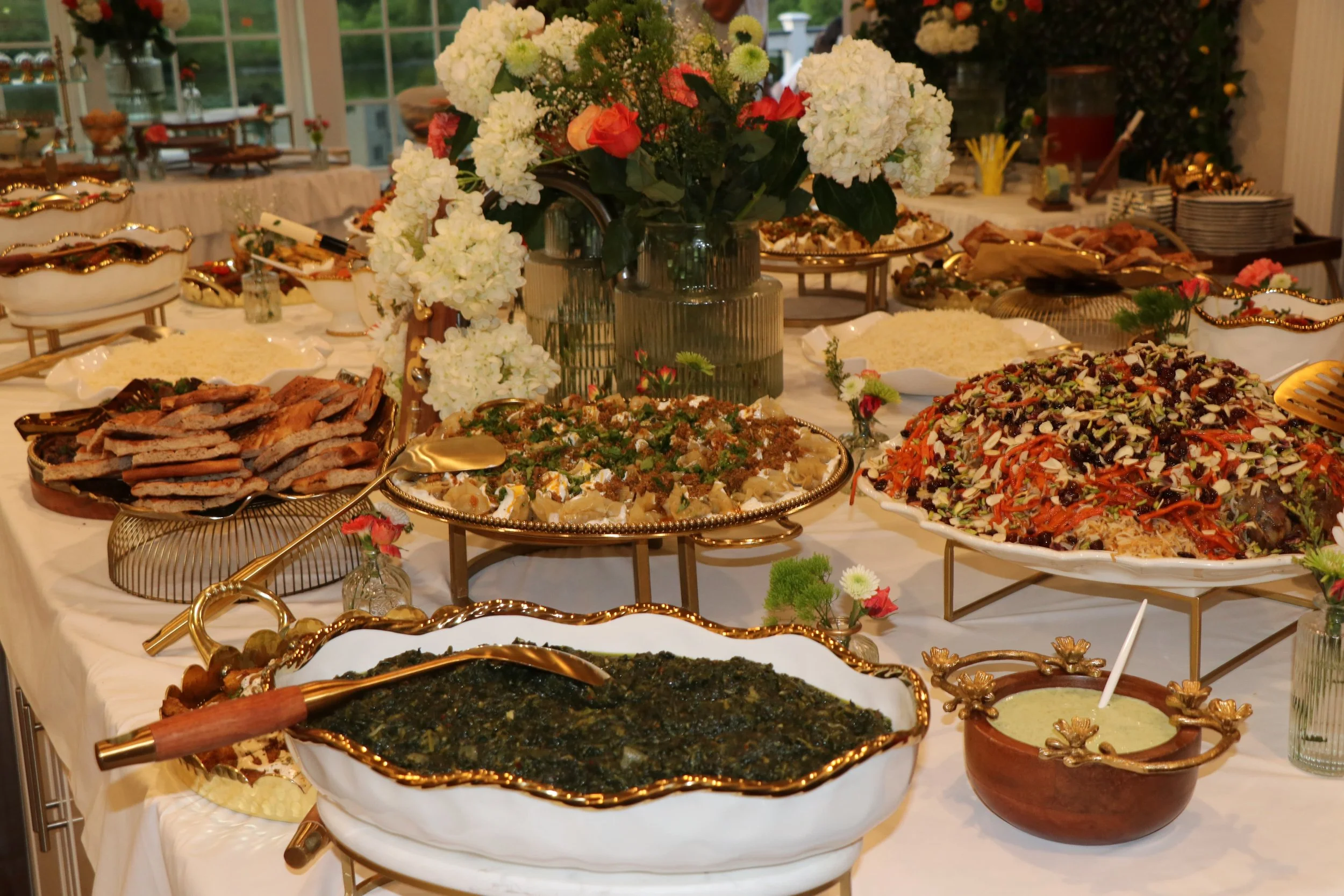 A buffet table with various dishes, including a large salad with shredded vegetables, a dish with greens, a bowl of creamy sauce, sliced meats, and other assorted foods, decorated with flowers.