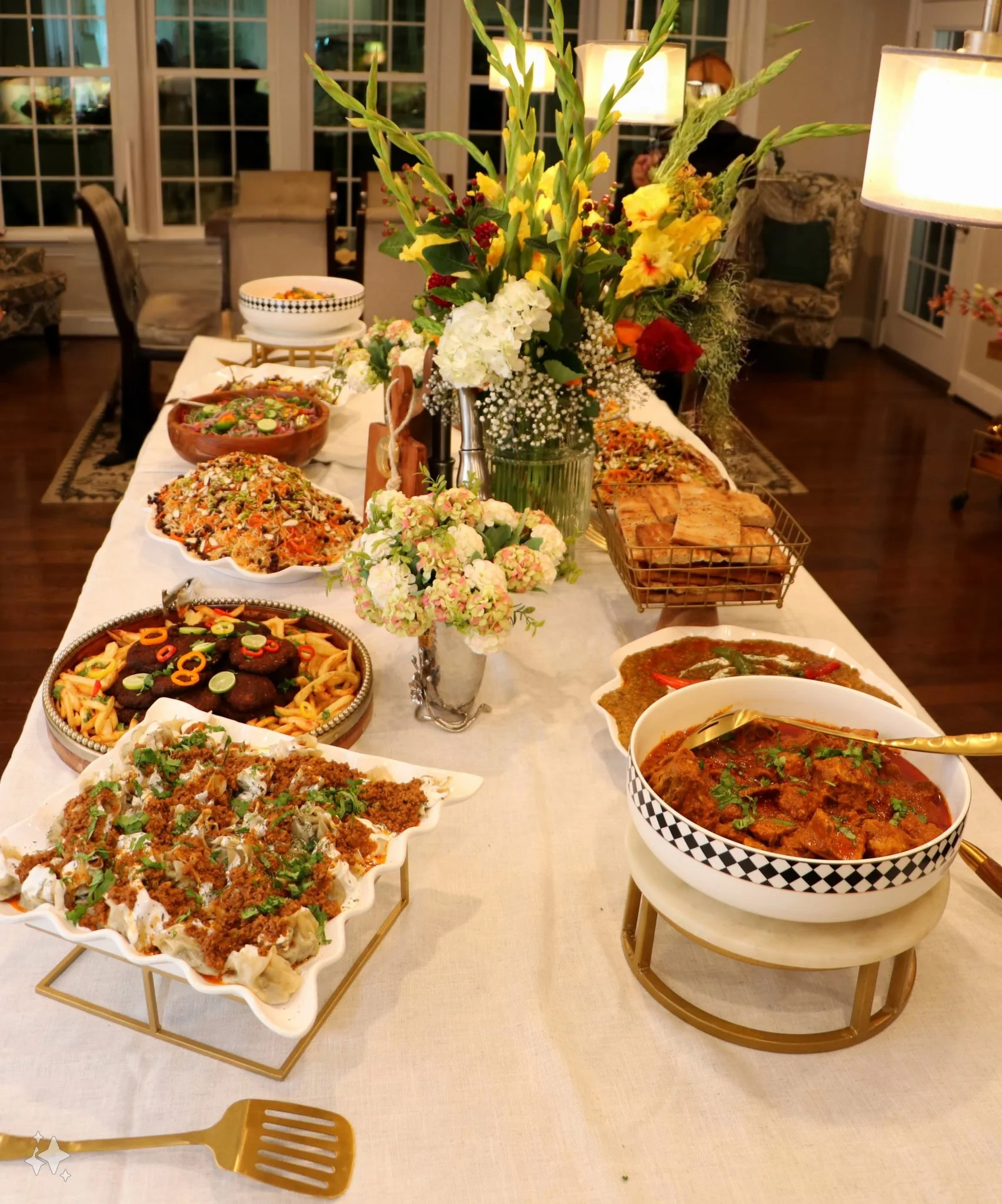 A table with various dishes of food including pasta, curry, salads, and bread, decorated with flowers, in a well-lit dining room.
