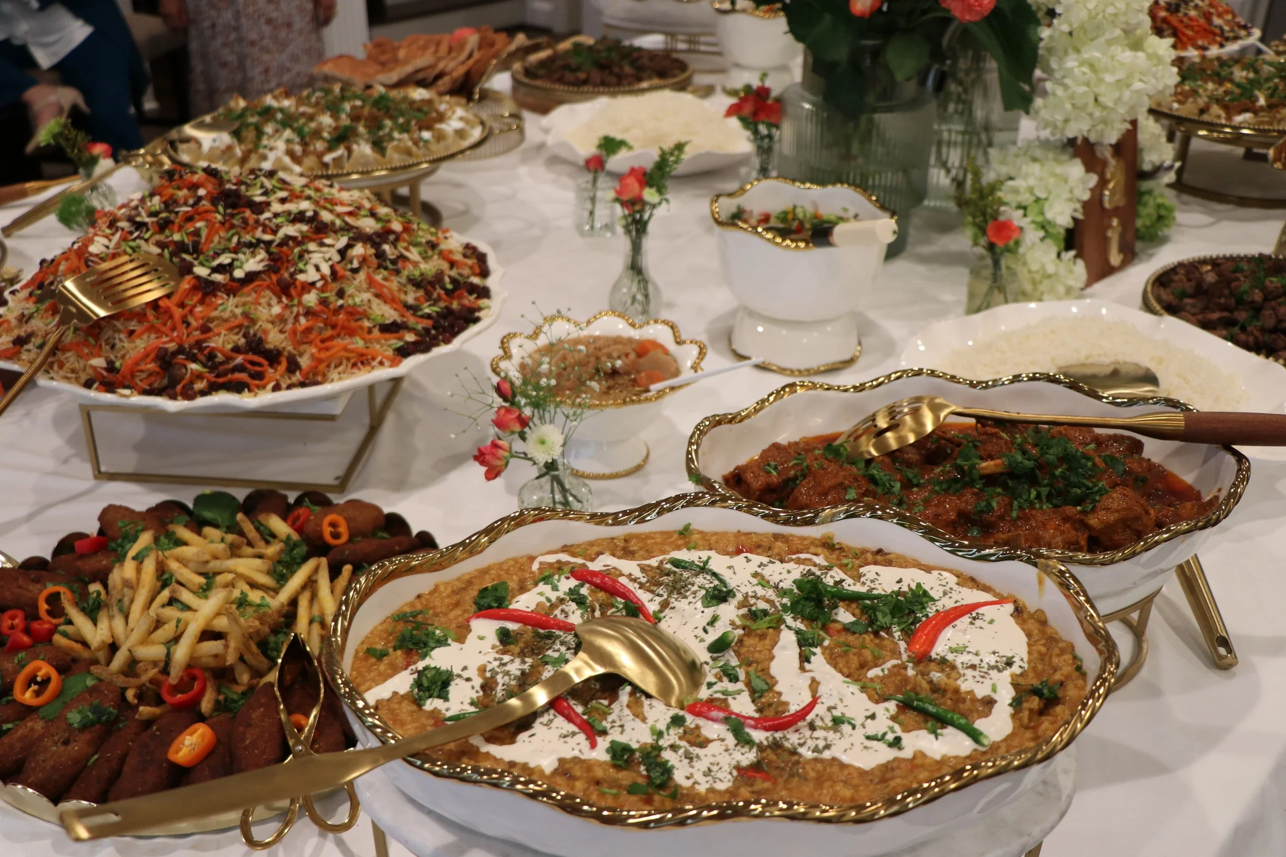 A buffet table with various dishes, including salads, meats, rice, and vegetables, decorated with flowers and gold-trimmed serving ware.