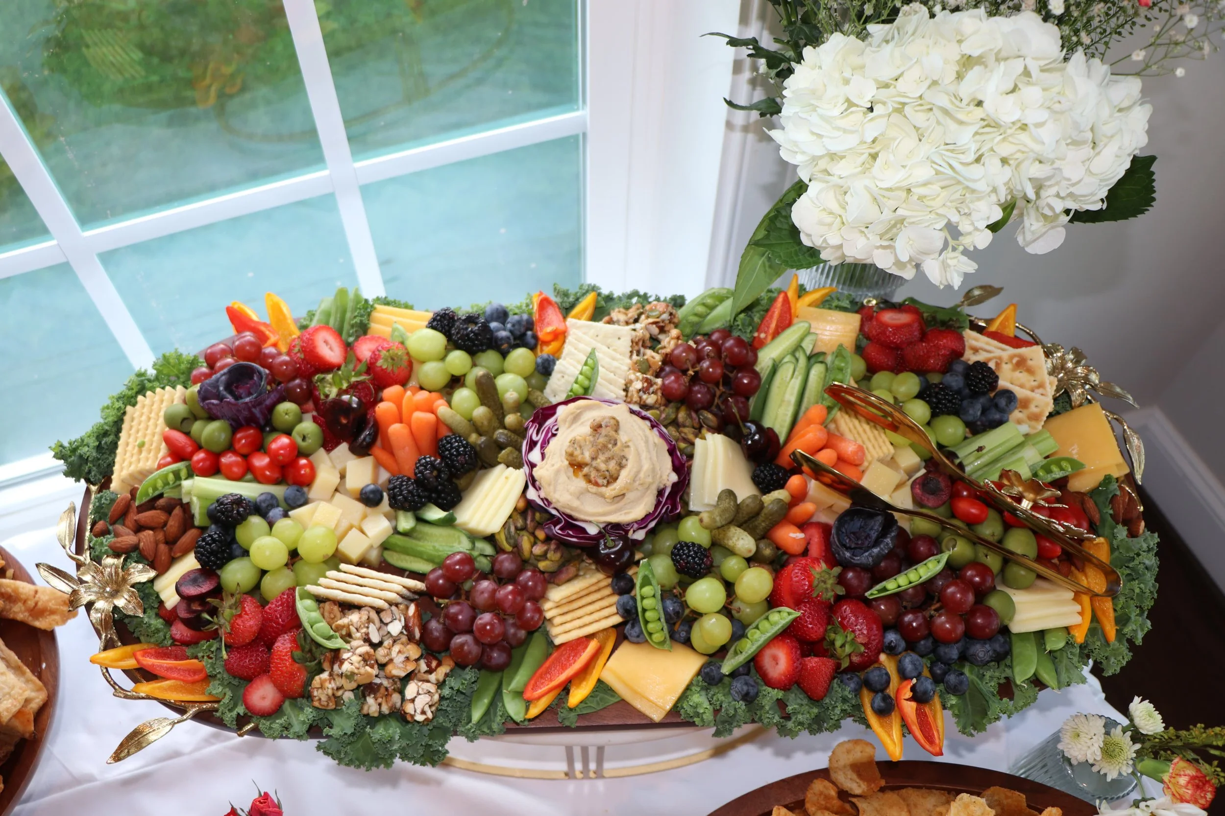 Large fruit and cheese platter with grapes, strawberries, blackberries, blueberries, crackers, and a floral centerpiece in white hydrangeas.