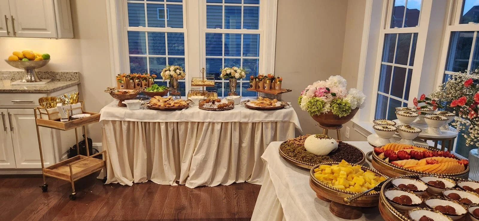 A table with assorted desserts, fruit, and flowers in a well-lit room with large windows.