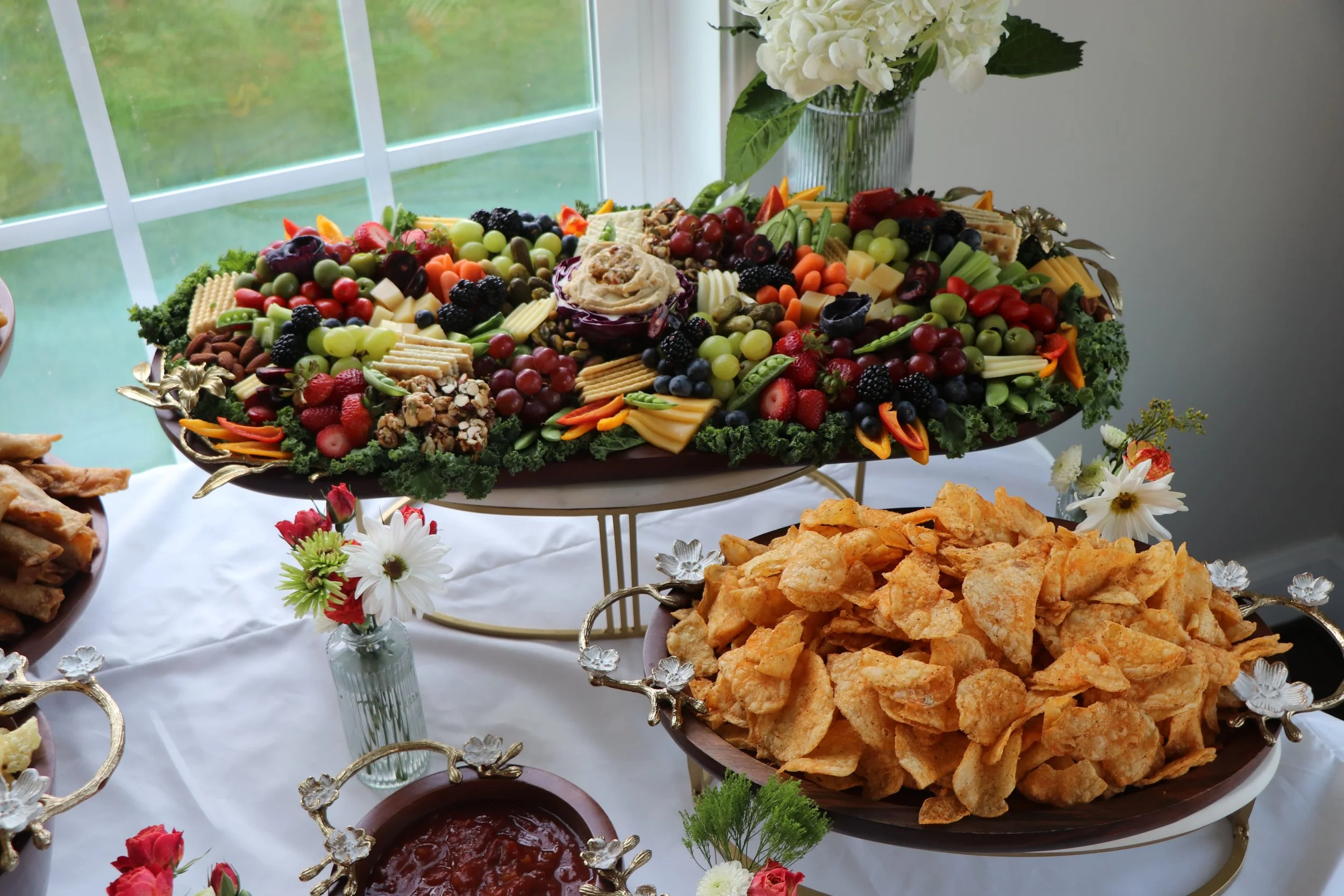 A large fruit and cheese platter with grapes, strawberries, blackberries, and assorted cheese slices, on a table with decorative flowers, beside a bowl of chips and a dish of dip.