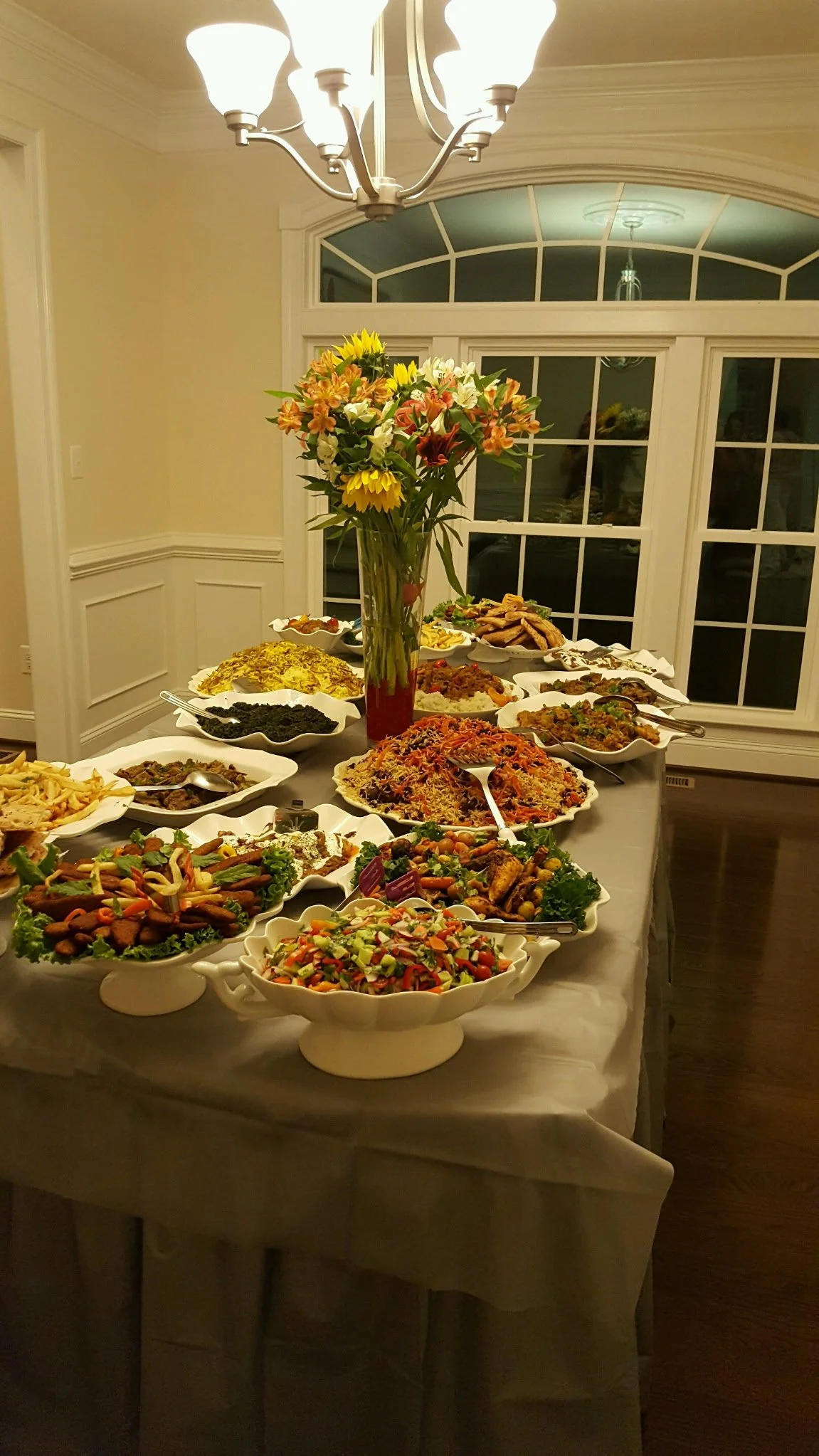 A buffet-style table with various dishes and a large floral centerpiece, located in a dining room with a large window and a chandelier overhead.