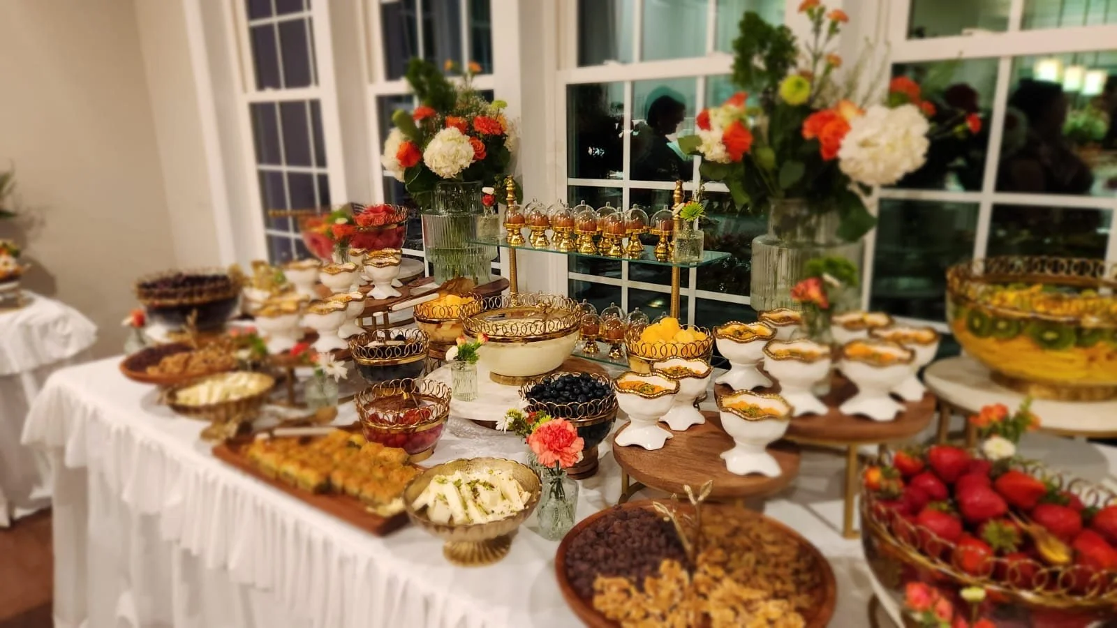 A dessert buffet table with various fruits, cakes, and pastries, decorated with flowers and gold accents, set in a well-lit room with large windows and white curtains.