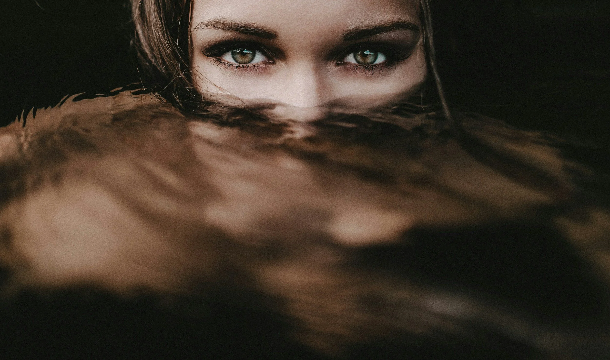 A close-up of a woman's face with striking green eyes, partially submerged in dark water with her hair blending into the water surface.