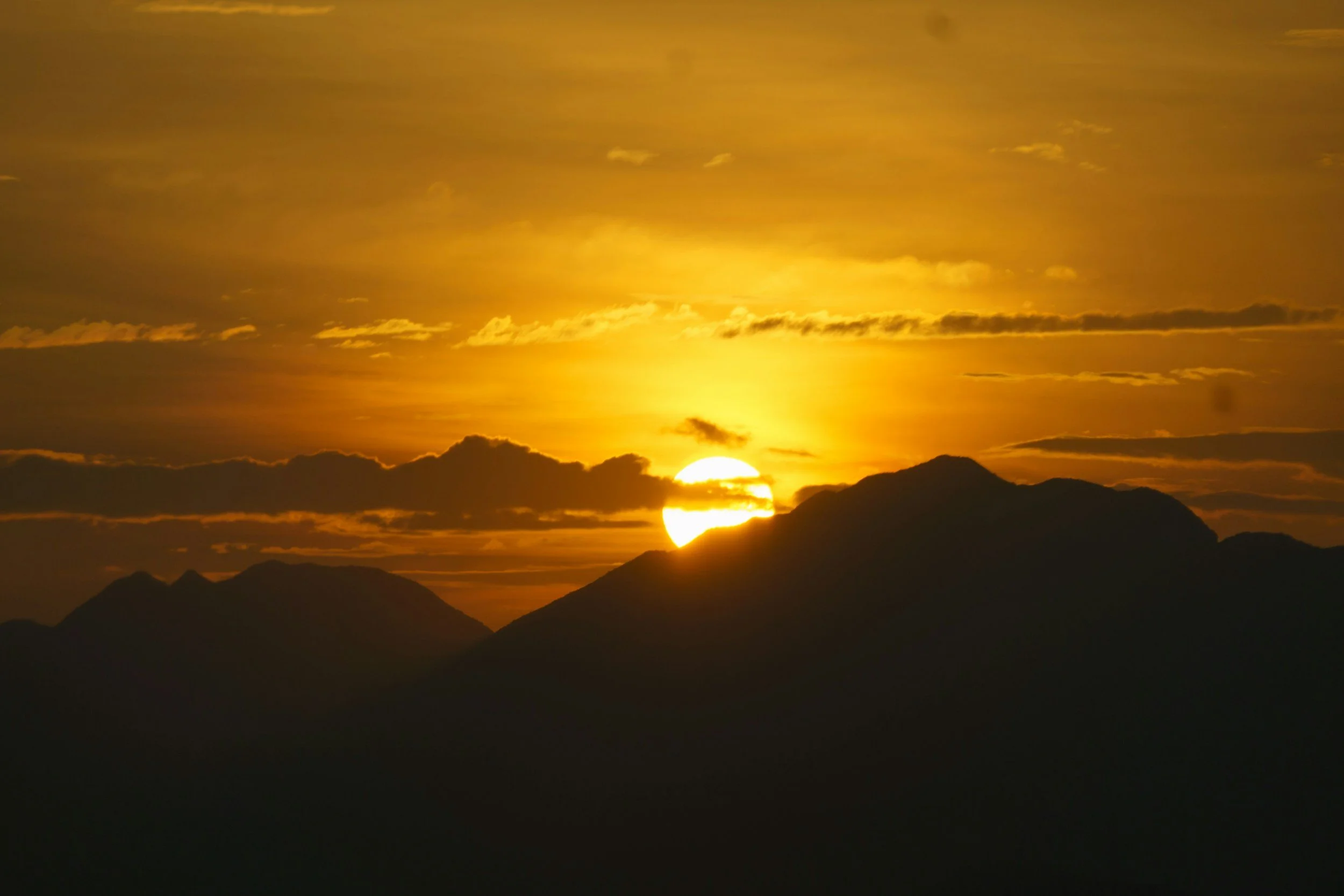 Sun setting behind silhouette of mountain range with orange and yellow sky and clouds.