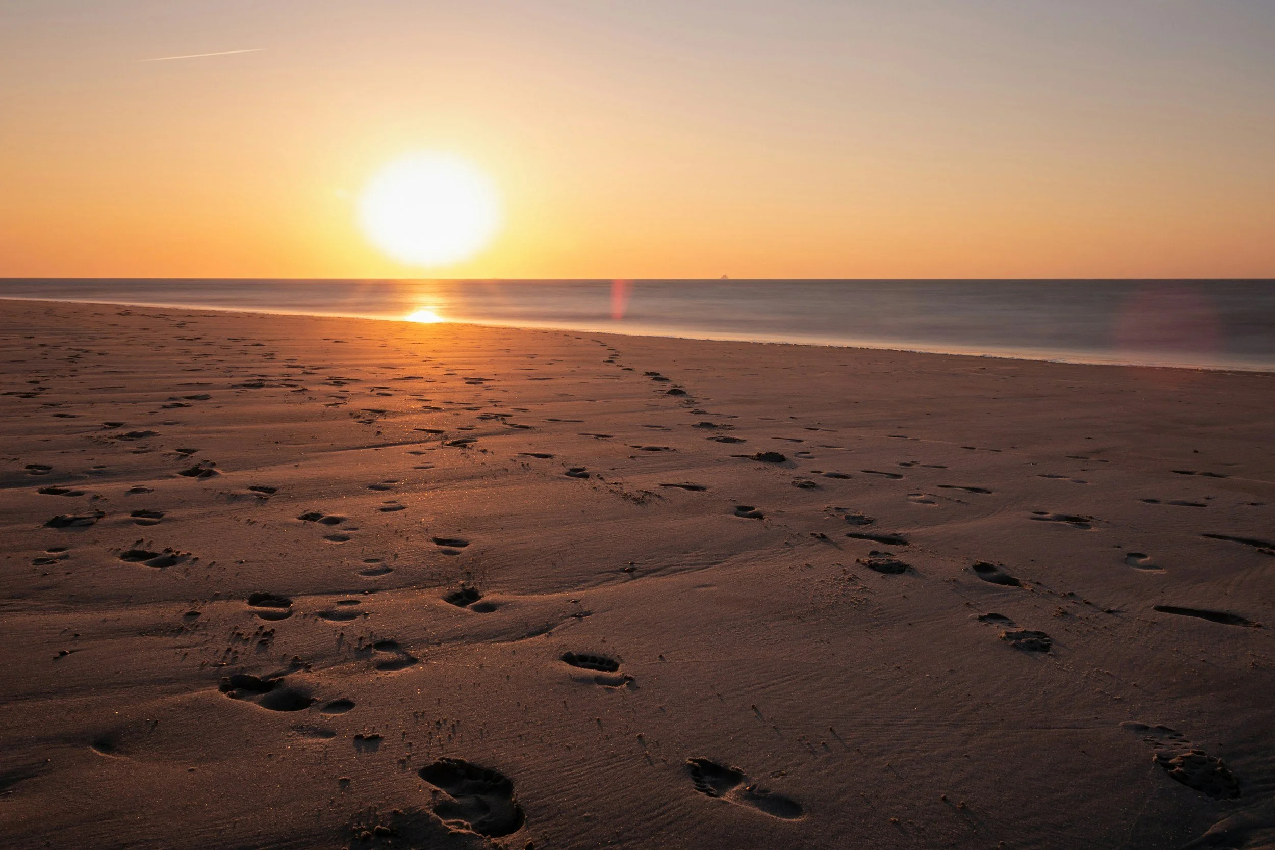 Footprints in the sand on a beach at sunset with the ocean and a ship in the distance.