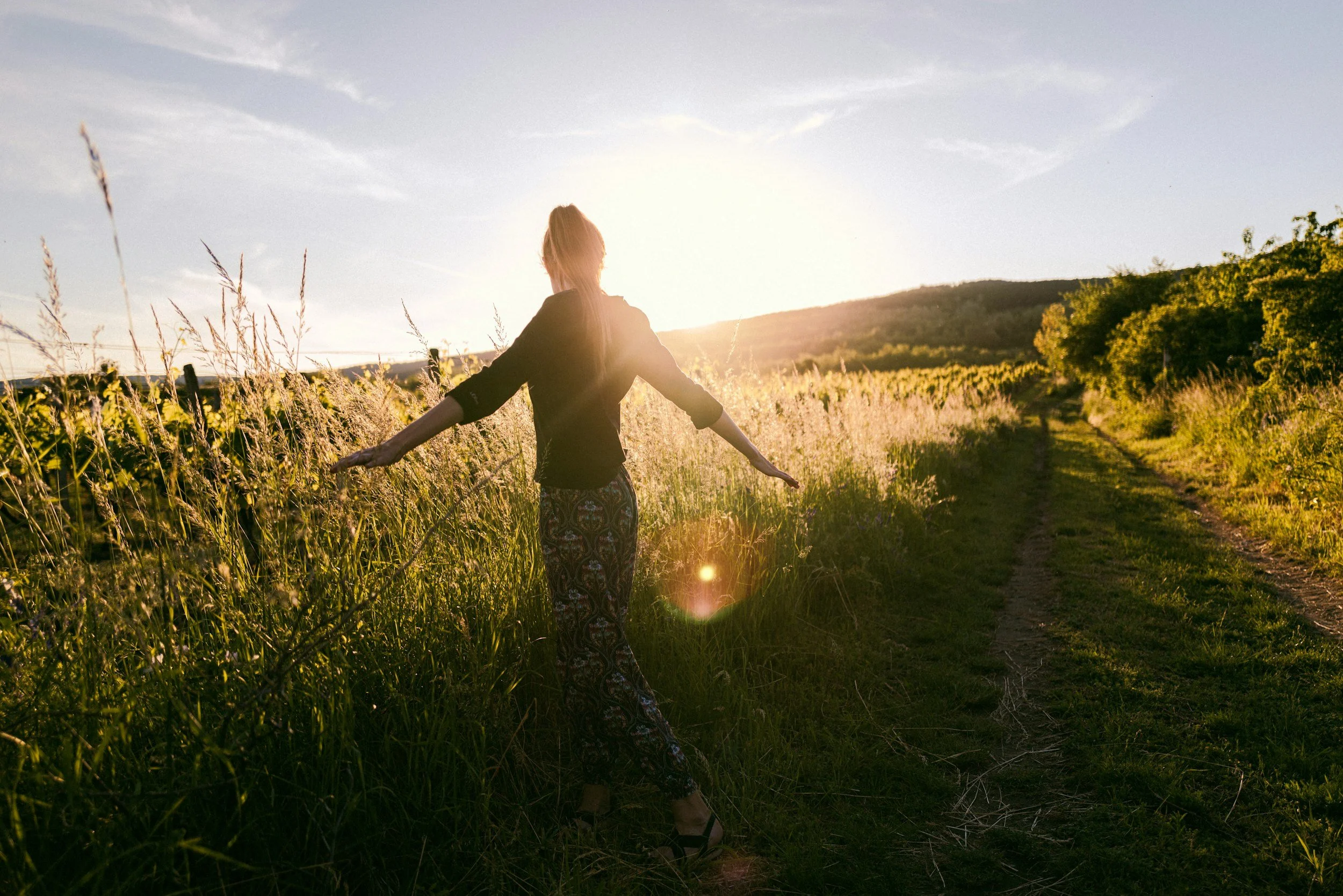 A woman standing on a grassy path in a field during sunset, with her arms outstretched and facing away from the camera.