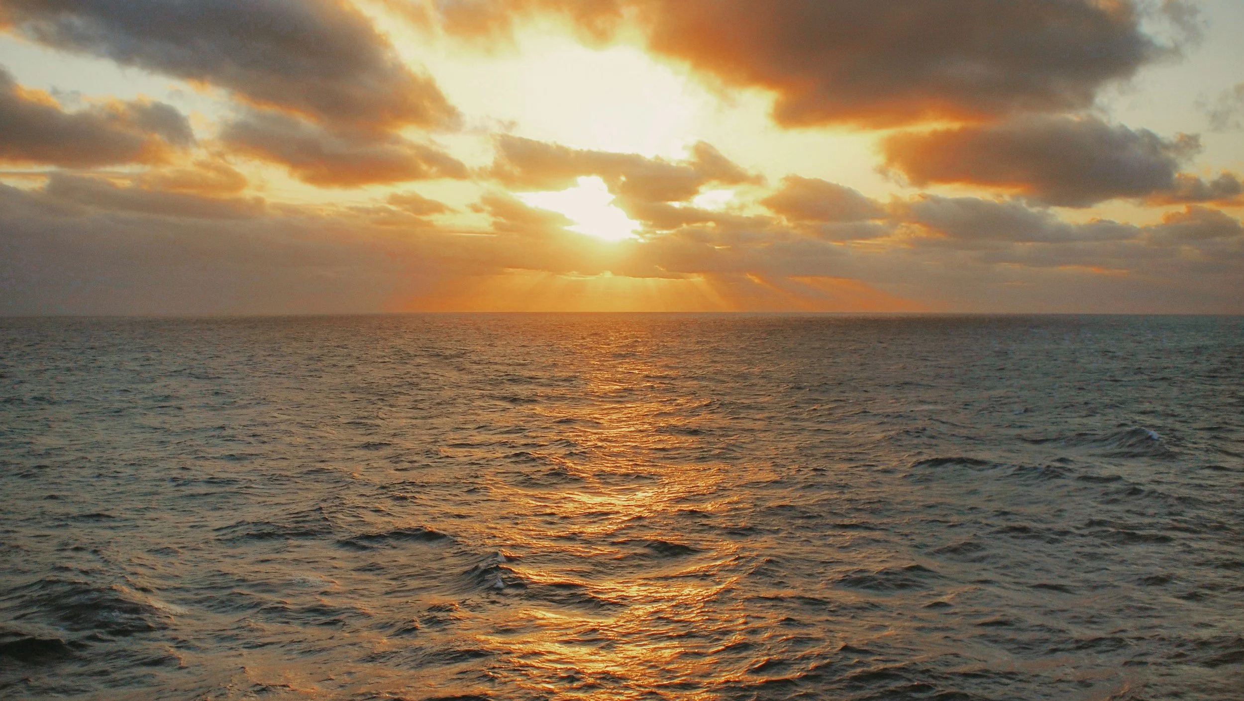 Sunset over the ocean with clouds in the sky and sunlight reflecting on the water.