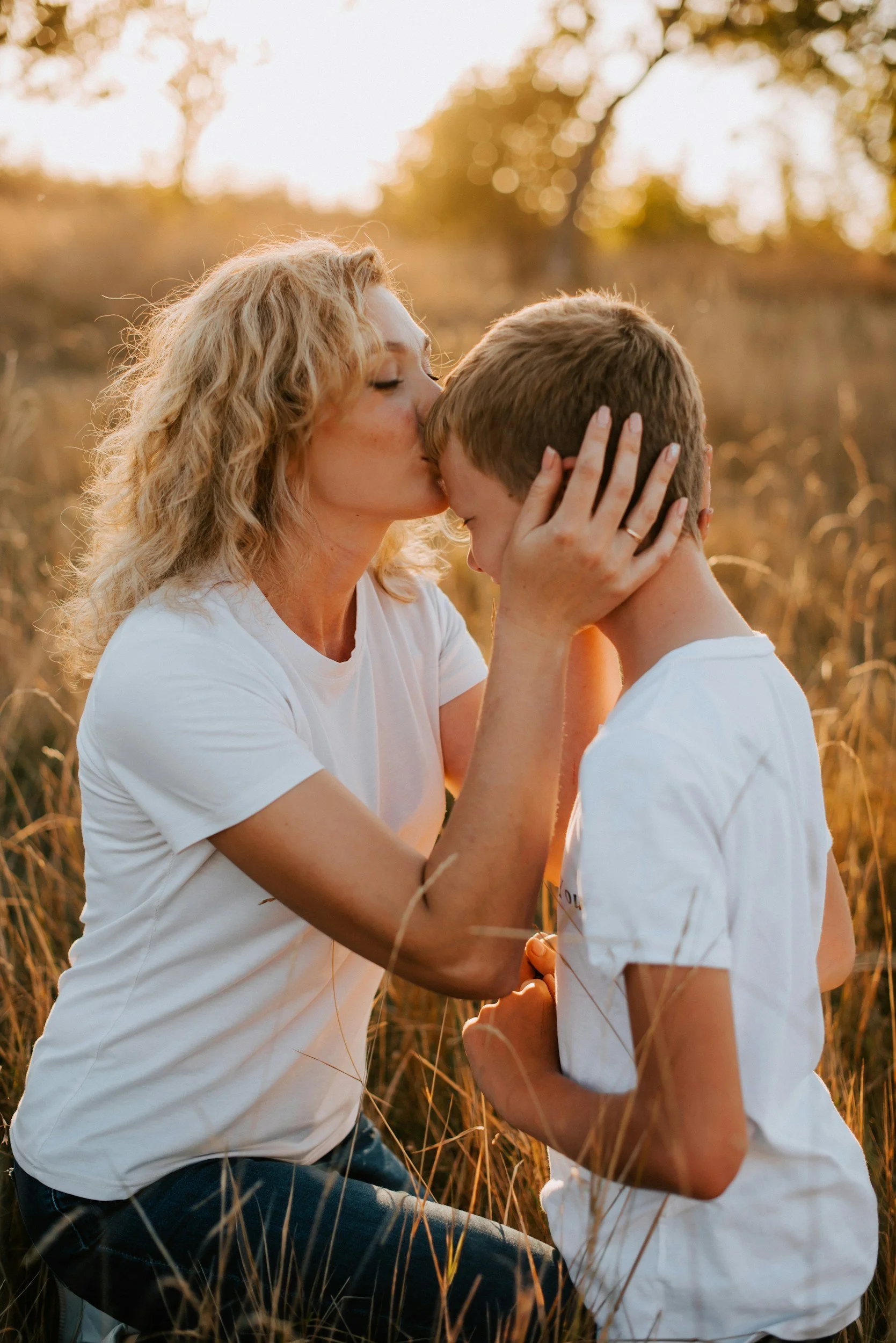 A woman with curly blonde hair wearing a white t-shirt kisses a boy on the forehead in a field during sunset, holding his face tenderly.