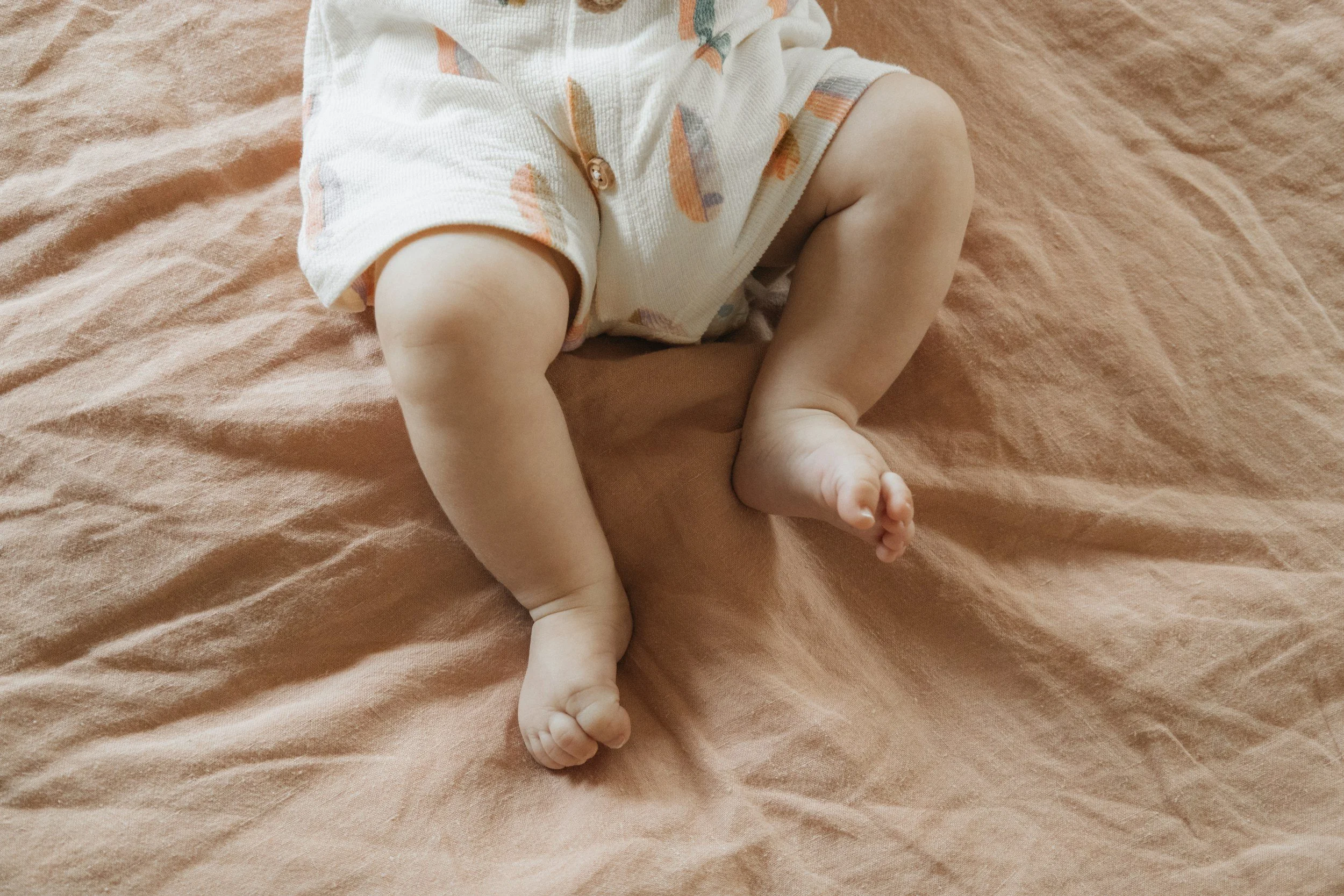 Close-up of a baby's legs and feet lying on a soft, beige blanket, wearing a light-colored outfit with multicolored patterns.