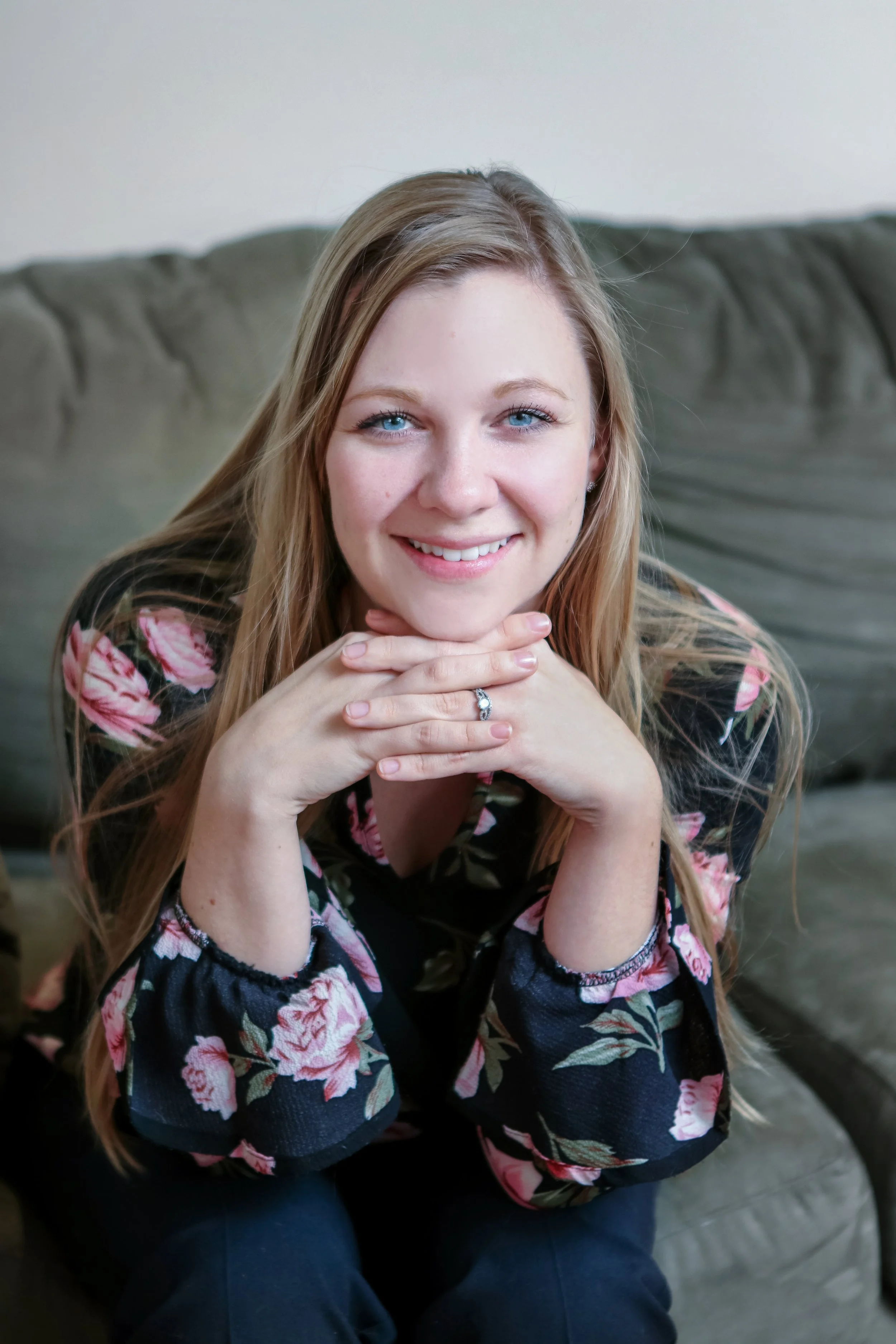 A young woman with long blonde hair, blue eyes, and a bright smile, sitting on a couch with hands clasped under her chin, wearing a floral-patterned black top.